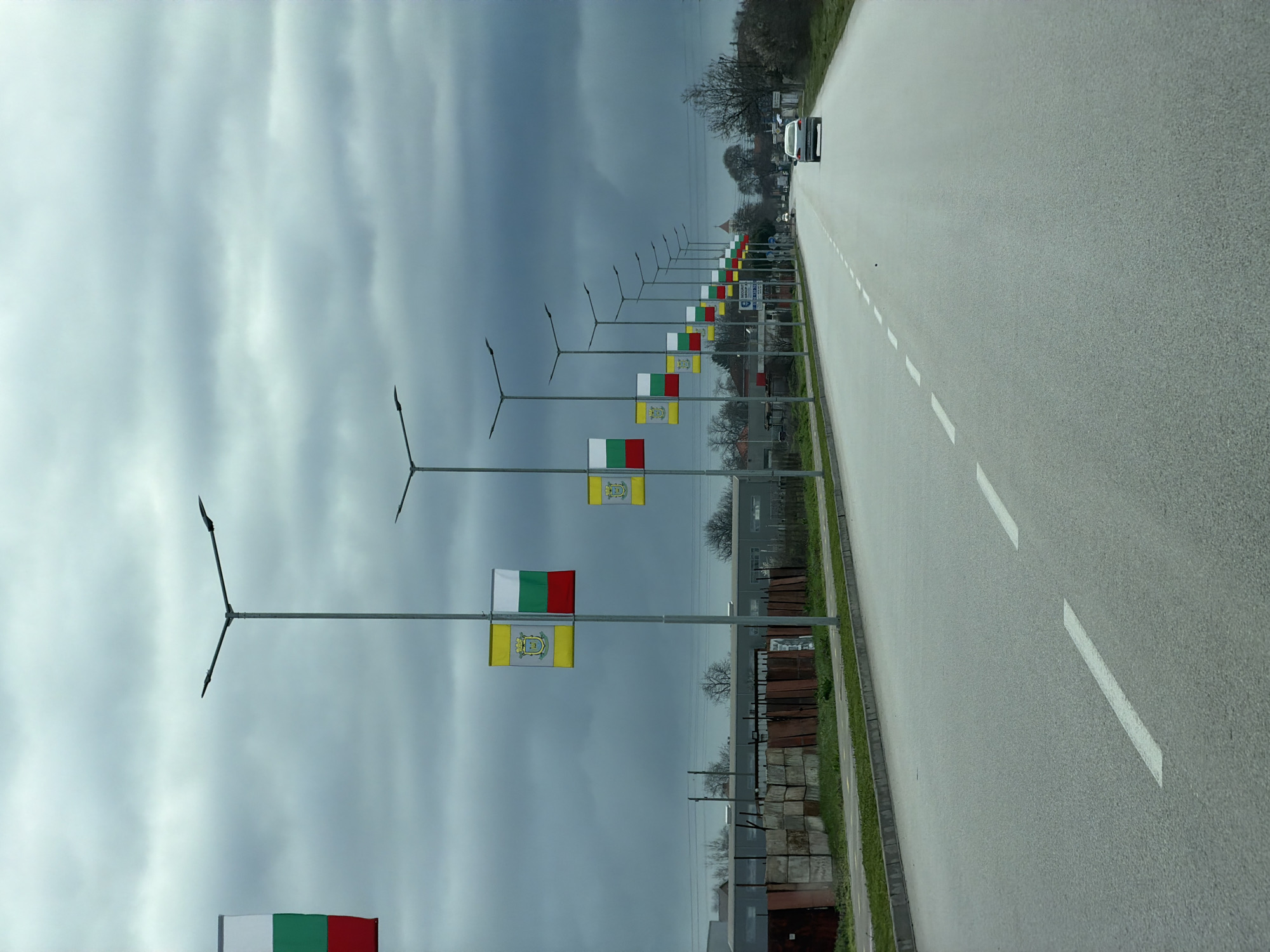 The image shows a road in Rakovski, Bulgaria lined with alternating Bulgarian national flags (white, green, red) and the flag of Rakovski (yellow with coat of arms). The flags are mounted on street light poles. A white car is driving on the road. The sky is overcast. There is a grassy verge between the road and a fence/wall, with a mix of buildings and construction materials visible behind the fence. The road has white dashed lane markings.