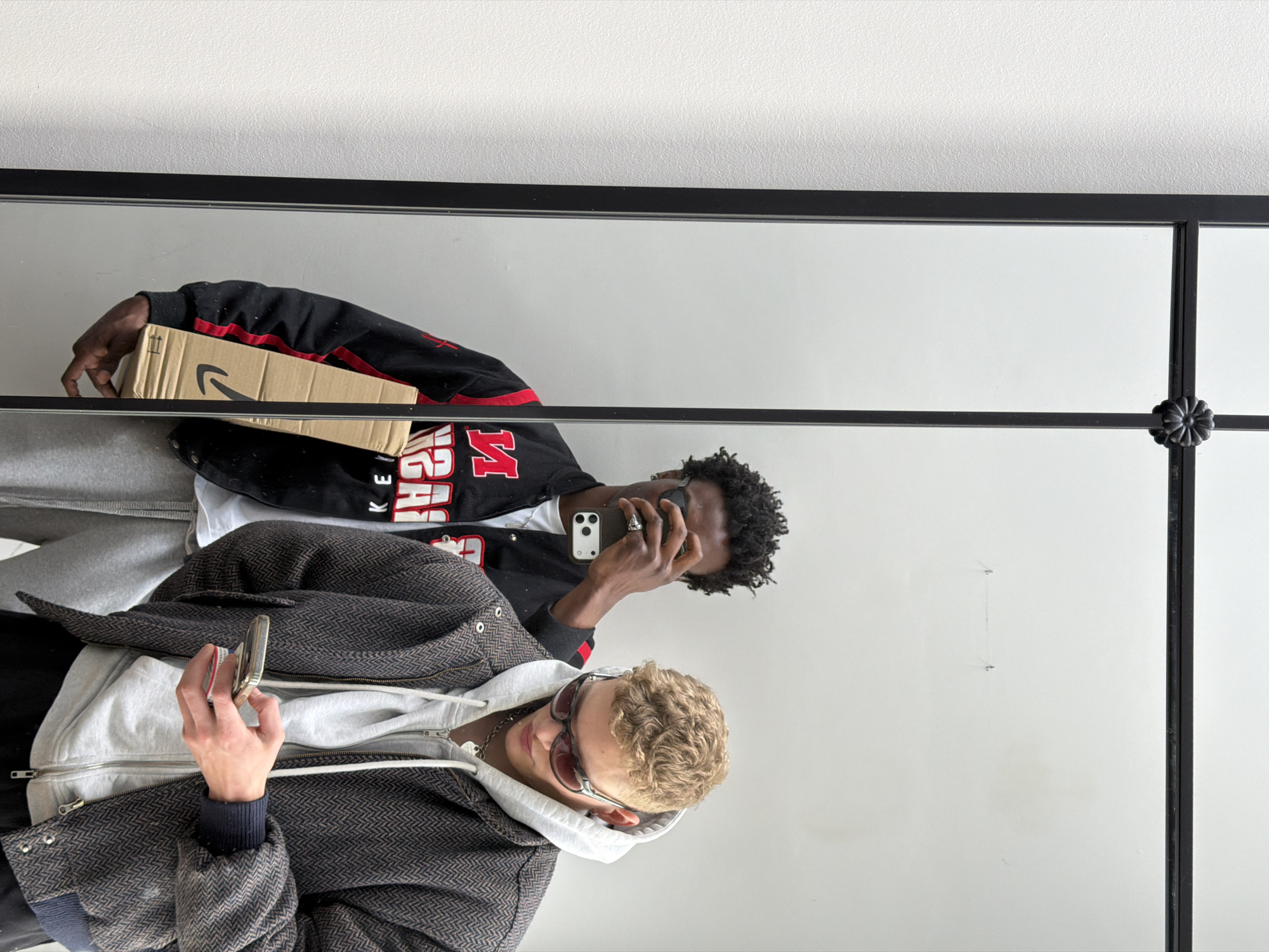A mirror selfie shows two individuals. One, with blond curly hair, wears a grey hoodie under a herringbone jacket, holding a phone. The other, of African descent with curly hair, wears a black and red jacket and holds an Amazon package, using his phone. The background consists of a white wall. The image does not depict elements that suggest the location is London, United Kingdom.