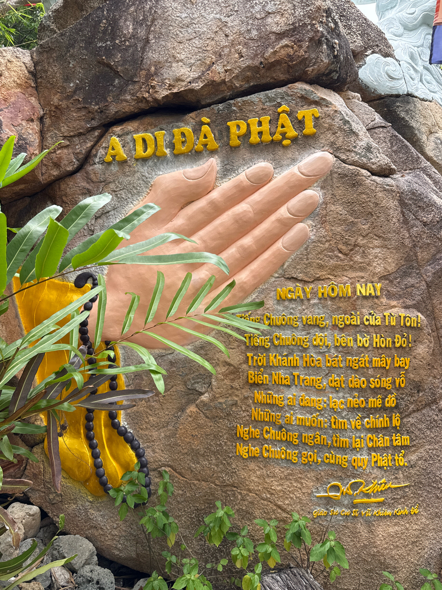 The image shows a relief carved into a large rock. The relief depicts two hands pressed together in a prayerful gesture, with the words "A DI DA PHAT" in large yellow letters above the hands. Below the hands, there is a poem written in Vietnamese, also in yellow letters. The poem begins with "NGAY HOM NAY" and continues to describe the beauty of Nha Trang and a call to seek inner peace. A yellow robe and a black mala are draped on the left side of the relief, partially obscuring the rock. Green foliage frames the lower left and right portions of the image. The rock itself appears to be part of a natural formation, possibly a cliff face. Based on the text, the relief is likely located in or near Nha Trang, Vietnam, and commemorates or refers to Buddhist teachings.