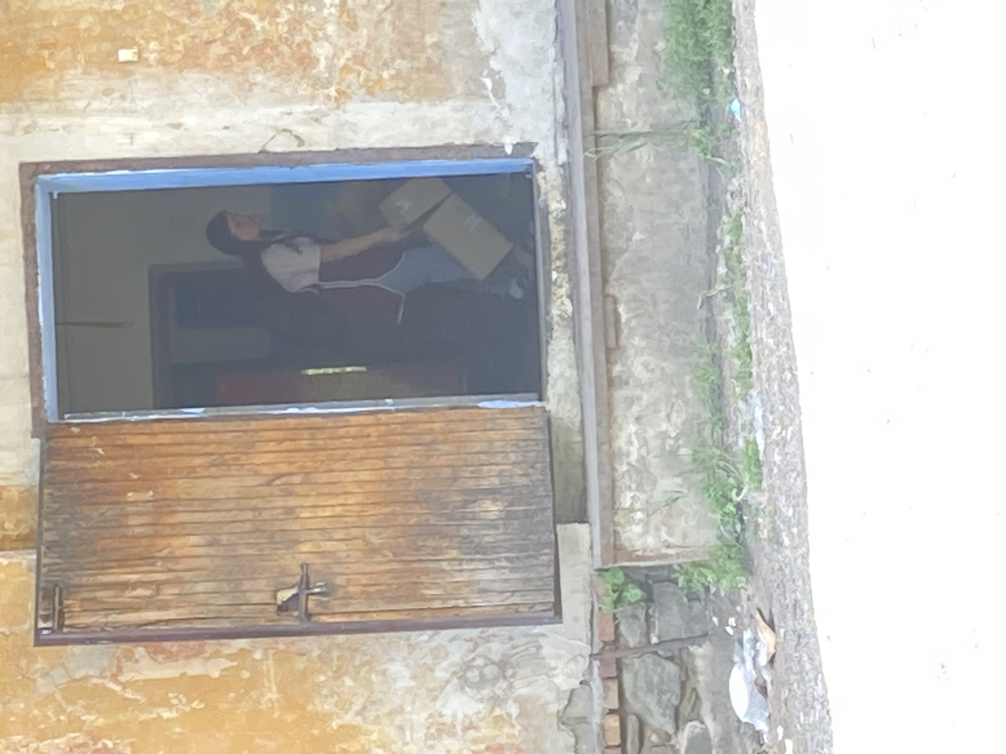 A person, likely female, with long dark hair and wearing a maroon apron over a white shirt and blue pants, exits a building through a wooden door. The person is carrying two cardboard boxes. The building has peeling orange and white paint on the exterior walls and a blue door frame. The ground in front of the building is gravelly, with some debris including a white plastic bag and a piece of orange paper. Green weeds grow near the foundation of the building. The scene is outdoors, under bright sunlight.