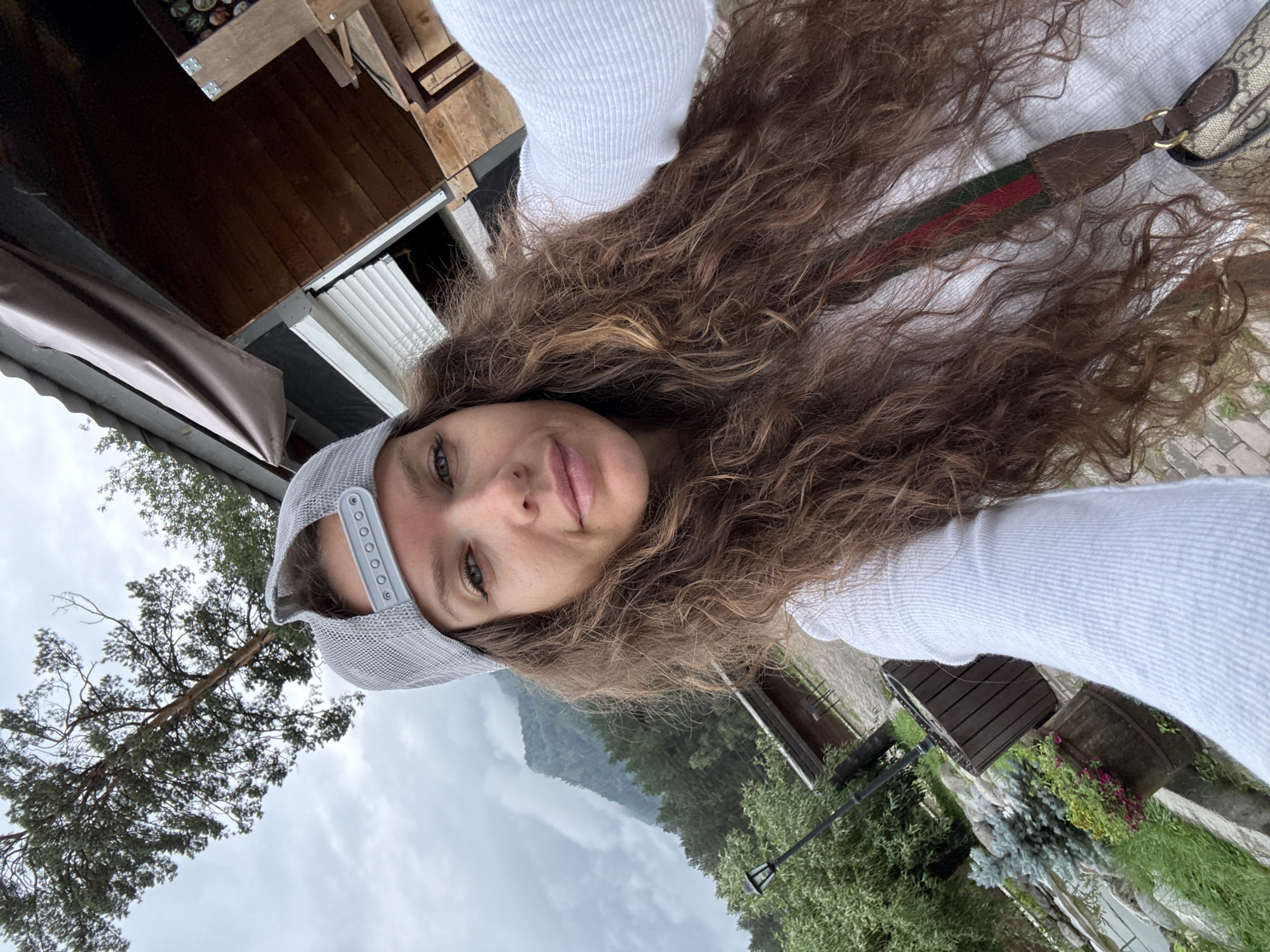 A selfie of a woman with long, curly brown hair and a gray baseball cap is taken outdoors. She is wearing a white long-sleeved shirt and a Gucci strap across her body. In the background, there is a wooden structure with a tarp, a tree, and a cloudy mountain landscape. The location is specified as the city of Barangol, Russia, implying a setting that is likely mountainous or in a rural area within Russia.