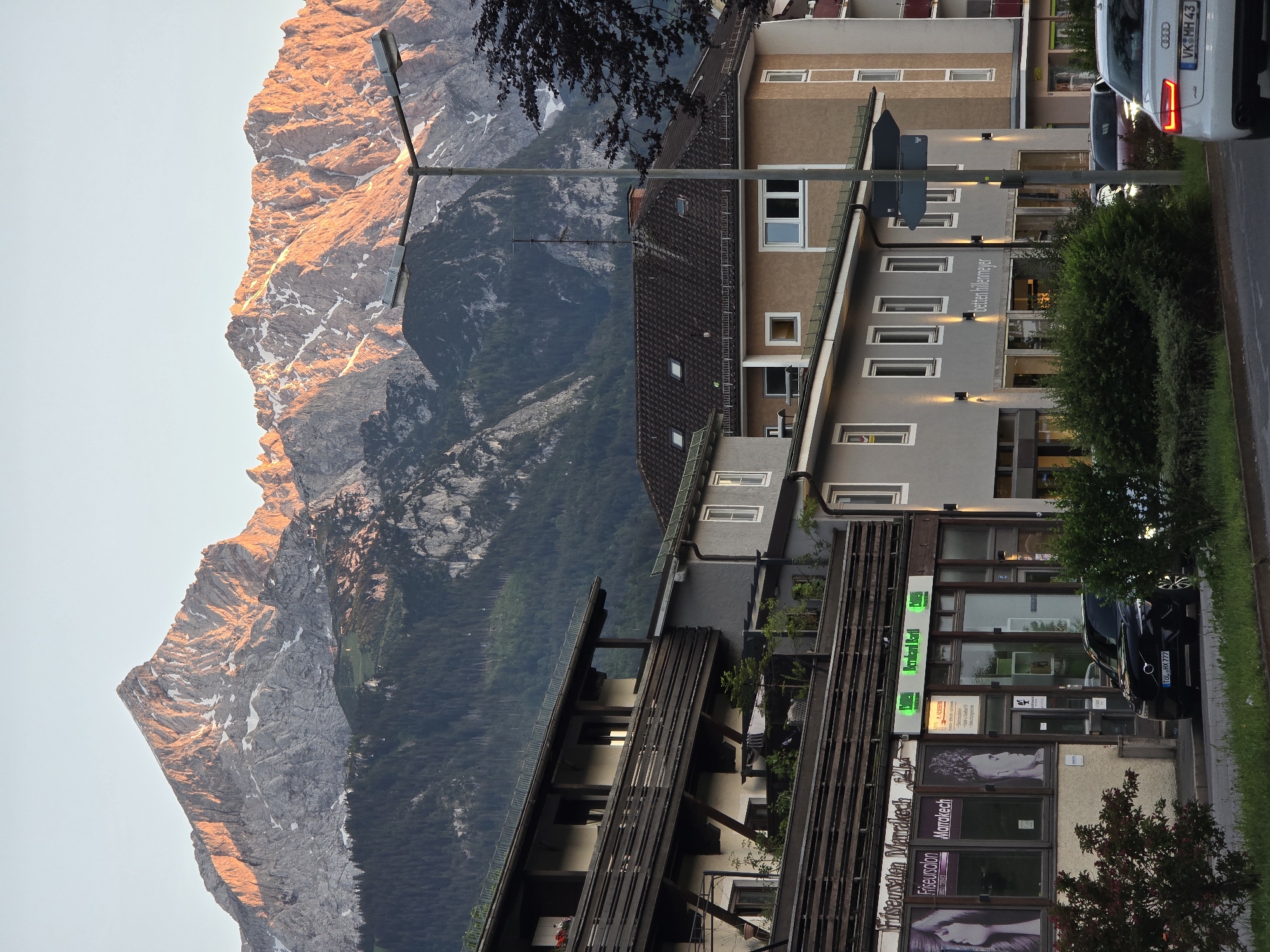 The image depicts a street scene in Garmisch-Partenkirchen, Germany, with a striking mountain backdrop. In the foreground, a street with parked cars is visible. A white Audi, with license plate VK MH 43, is in the right lane, its taillights illuminated. To its left, a black Volvo sedan, with license plate TOL HX 777, is parked.
On the right side of the street, several buildings are present. One building, painted a light brown, has multiple windows and a tiled roof. Adjacent to it, a narrower building with a gray facade features several lit exterior lights and signage that reads "betten hillenmeyer". Further to the left, a multi-story building with dark wooden balconies overlooks the street. The ground floor of this building houses businesses, including a hair salon named "Friseursalon Marrakech" and another establishment with green signage displaying "Bernard Bartl".
The background is dominated by a majestic mountain range, illuminated by the warm glow of the setting sun. The peaks are jagged and rocky, with patches of snow visible, and the slopes are covered in dense green forest. The sky is clear and a pale blue, transitioning to a warmer hue near the horizon. A street lamp with two fixtures stands prominently in the mid-ground, positioned between the buildings and the mountains.