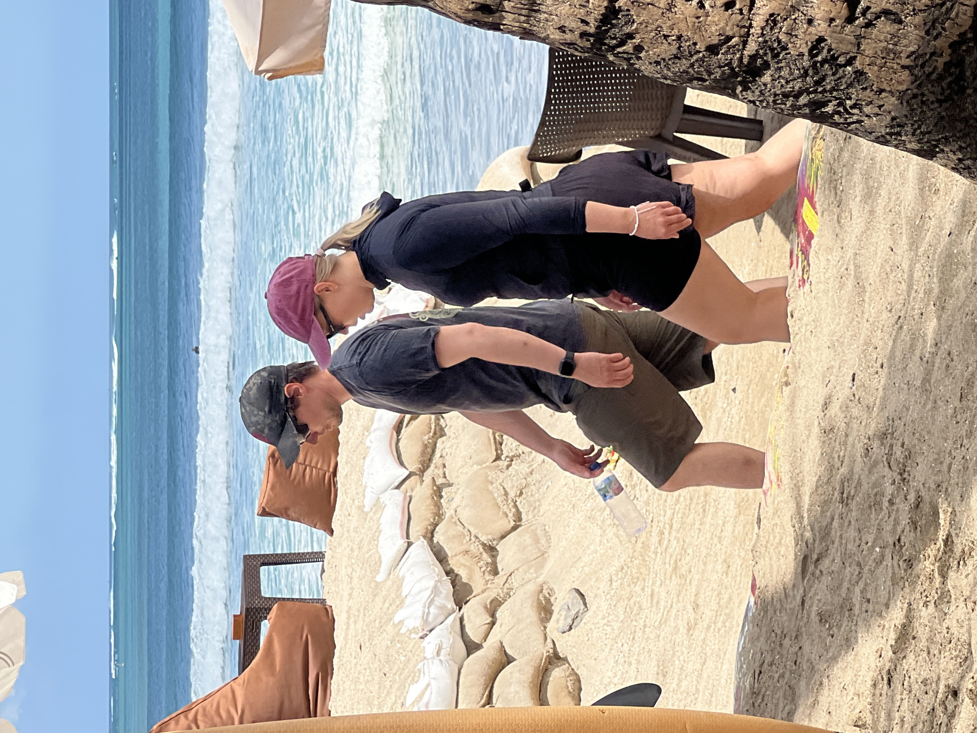 Two individuals are walking on a sandy beach in Kuta, Indonesia, with the ocean and waves visible in the background. The person on the left, a male, wears a dark grey t-shirt, dark green shorts, and a black baseball cap. He is holding a clear plastic water bottle in his right hand. The person on the right, a female, wears a black long-sleeved rashguard, black shorts, and a maroon baseball cap. She has blonde hair pulled back in a ponytail and wears a white bracelet on her left wrist. They are walking in the same direction, with the man slightly ahead. In the foreground, sandbags are piled up, possibly as a barrier, and there are large beanbag chairs and a small table to the left, suggesting a seating area. A palm tree trunk is visible on the far right. The water is turquoise with white waves breaking closer to shore. The sky is clear and blue.