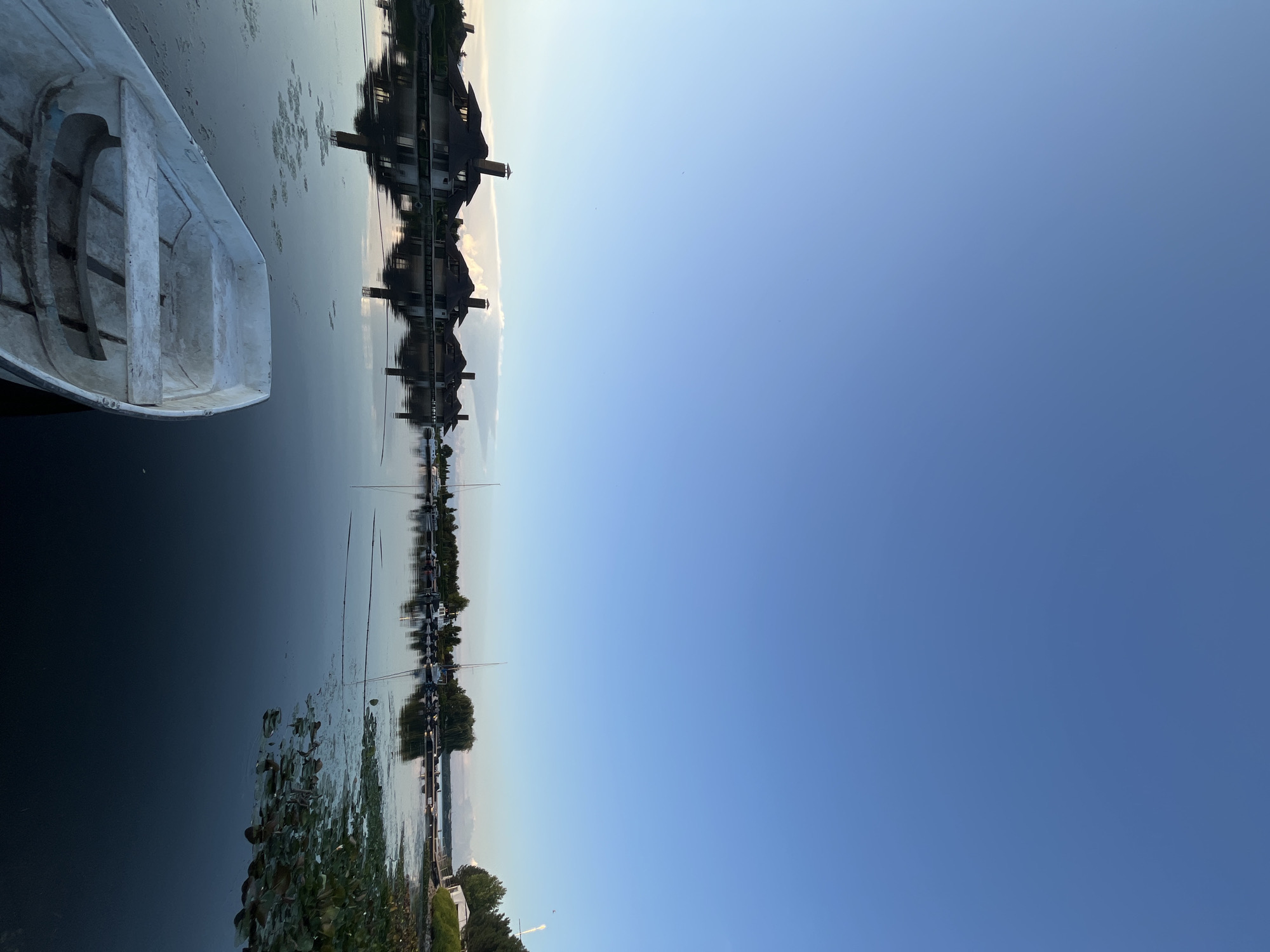 The image shows a tranquil scene on a body of water, likely a river or lake, with a clear blue sky overhead. In the foreground, a small, white rowboat rests on the water, its interior showing signs of wear. The middle ground features a row of houses with distinctive dark, pitched roofs and prominent chimneys, reflecting in the calm water. Beyond the houses, a marina is visible with several boats and yachts docked. The far background consists of trees and the horizon. The scene is likely located in the city of Dnipro, Ukraine, given the architectural style and the presence of a waterfront residential area with a marina. There are no people visible in the image. The overall impression is one of peaceful serenity in a waterside community.