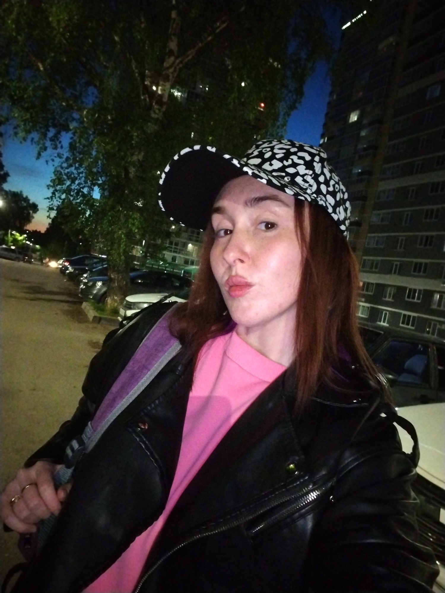 A woman is standing outside at night in Izhevsk, Russia. She is wearing a black leather jacket over a pink shirt and a black and white patterned baseball cap. She has auburn hair and is pursing her lips. A building with many windows is visible in the background, along with trees and parked cars. The overall lighting suggests it is dusk or night.
