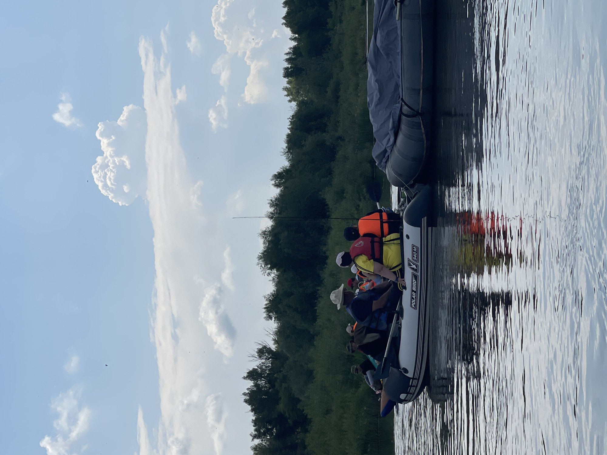 The image shows an inflatable boat with several people on board, navigating a body of water. The boat is white with black trim and has the brand "POLAR BIRD" and model "340 M" visible on its side. People are wearing life jackets in various colors, including yellow, orange, and blue. Some are wearing hats. The boat is moving away from the viewer, towards a dense treeline on the opposite bank of the water. The sky above is partially cloudy with a light blue hue. The water reflects the sky and the treeline. Another, larger inflatable object, covered with a gray tarp, is tied to the side of the main boat. The scene appears to be a recreational outing on a river or lake. The location is identified as Nezhinka, Russia, suggesting a natural environment.