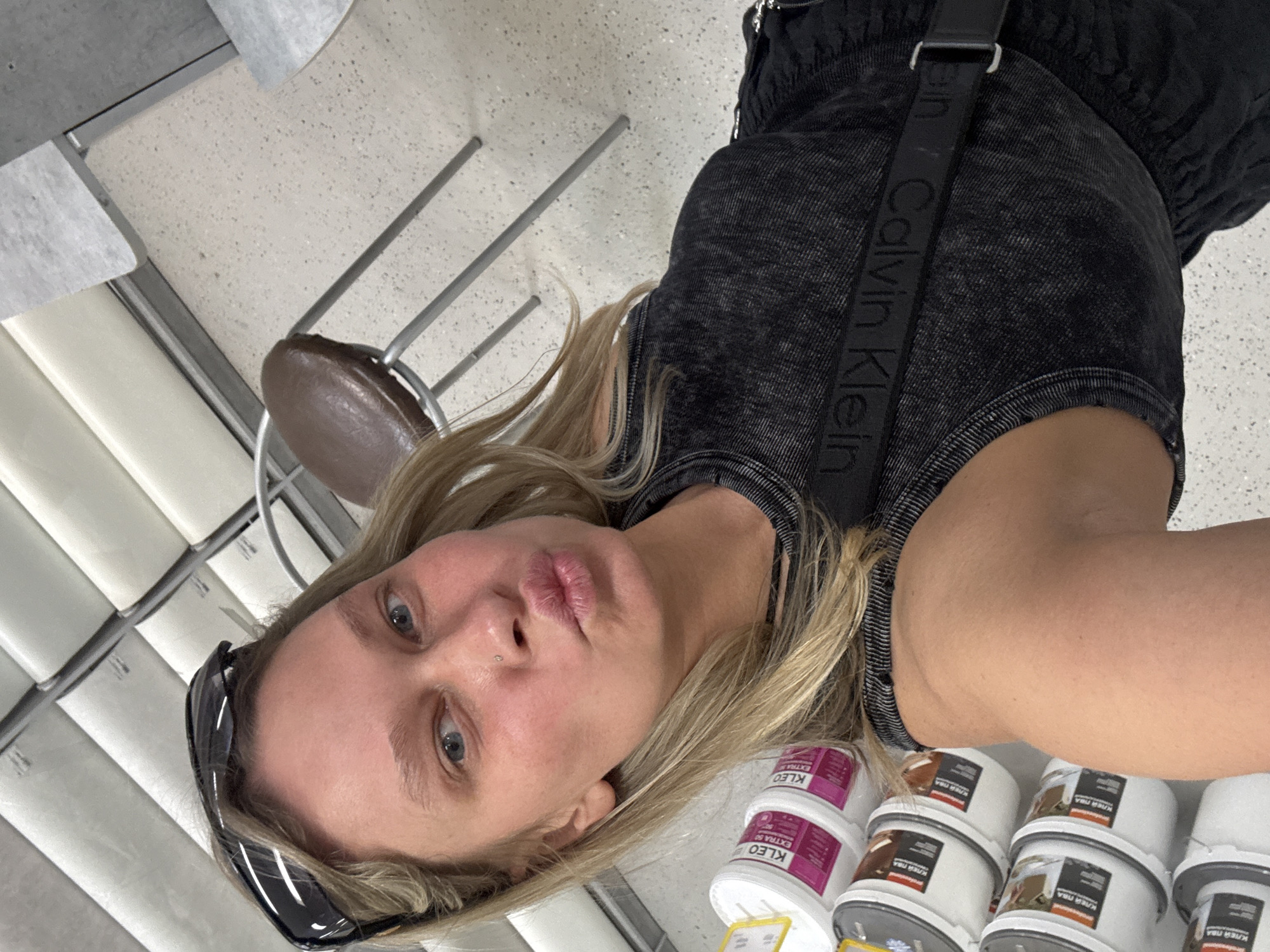 A selfie shows a blonde woman with sunglasses perched atop her head, positioned in what appears to be a hardware or paint store. She is wearing a black top with a Calvin Klein strap across her chest. The foreground shows shelves stocked with buckets of paste or glue with the label "KLEO EXTRA 50" and "Клей ПВА", a type of PVA glue in Russian. Behind her, the background features shelving and a chair, suggesting she is in a retail setting, possibly in the city of Perm, Russia.