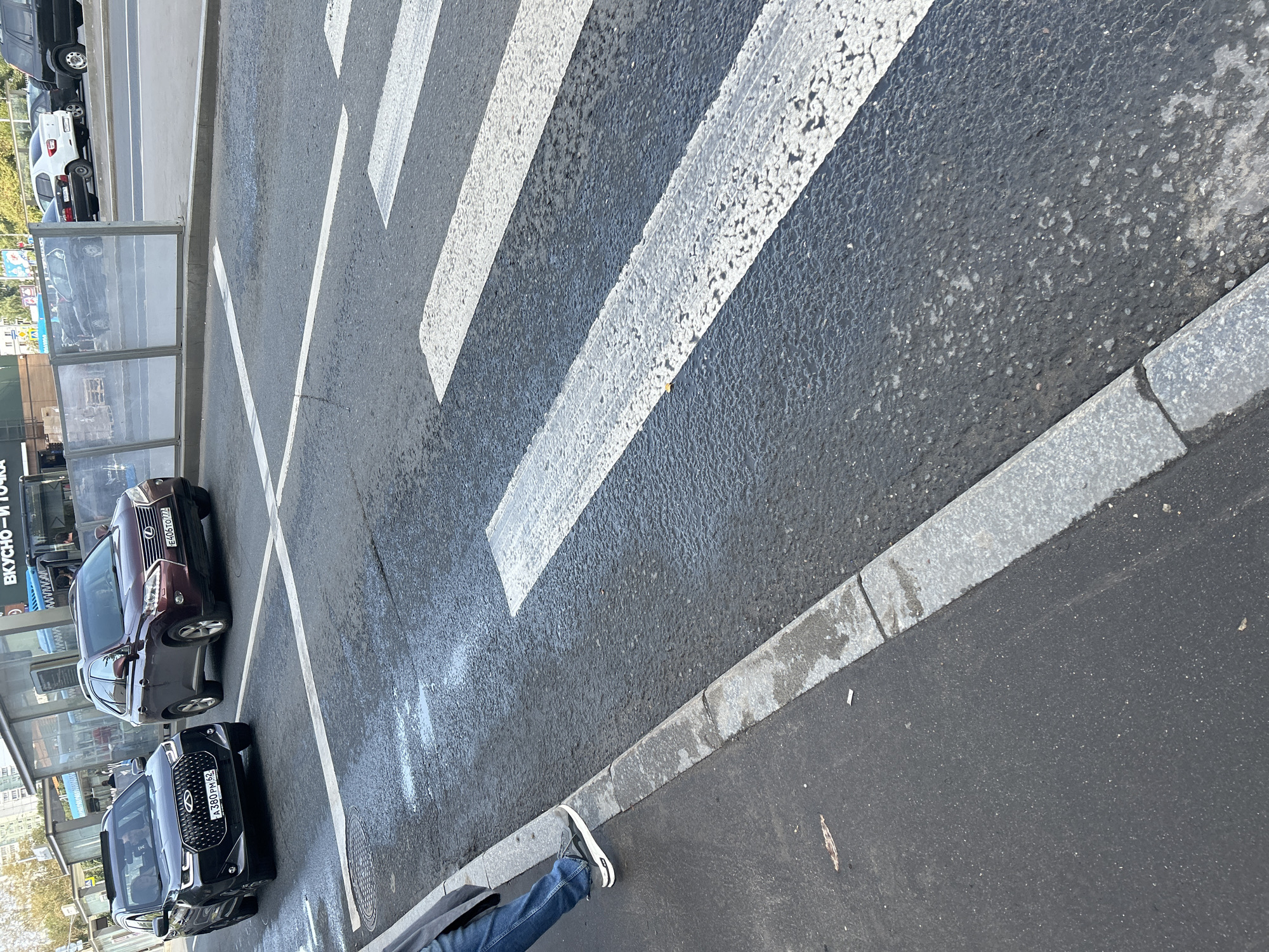 The image shows a street scene in Moscow, Russia, with a pedestrian crossing in the foreground. A dark gray SUV and a maroon Lexus SUV are visible on the road, stopped at or near the crossing. The Lexus has a license plate that reads "E406TO 777". The dark gray SUV has a license plate "A 380 PM 62". A person's legs and feet, wearing jeans and sneakers, are visible on the sidewalk, indicating they are either waiting to cross or have just crossed. The road appears wet in places, possibly from recent rain or cleaning. In the background, there are buildings with storefronts and some parked vehicles. The overall environment suggests an urban setting.