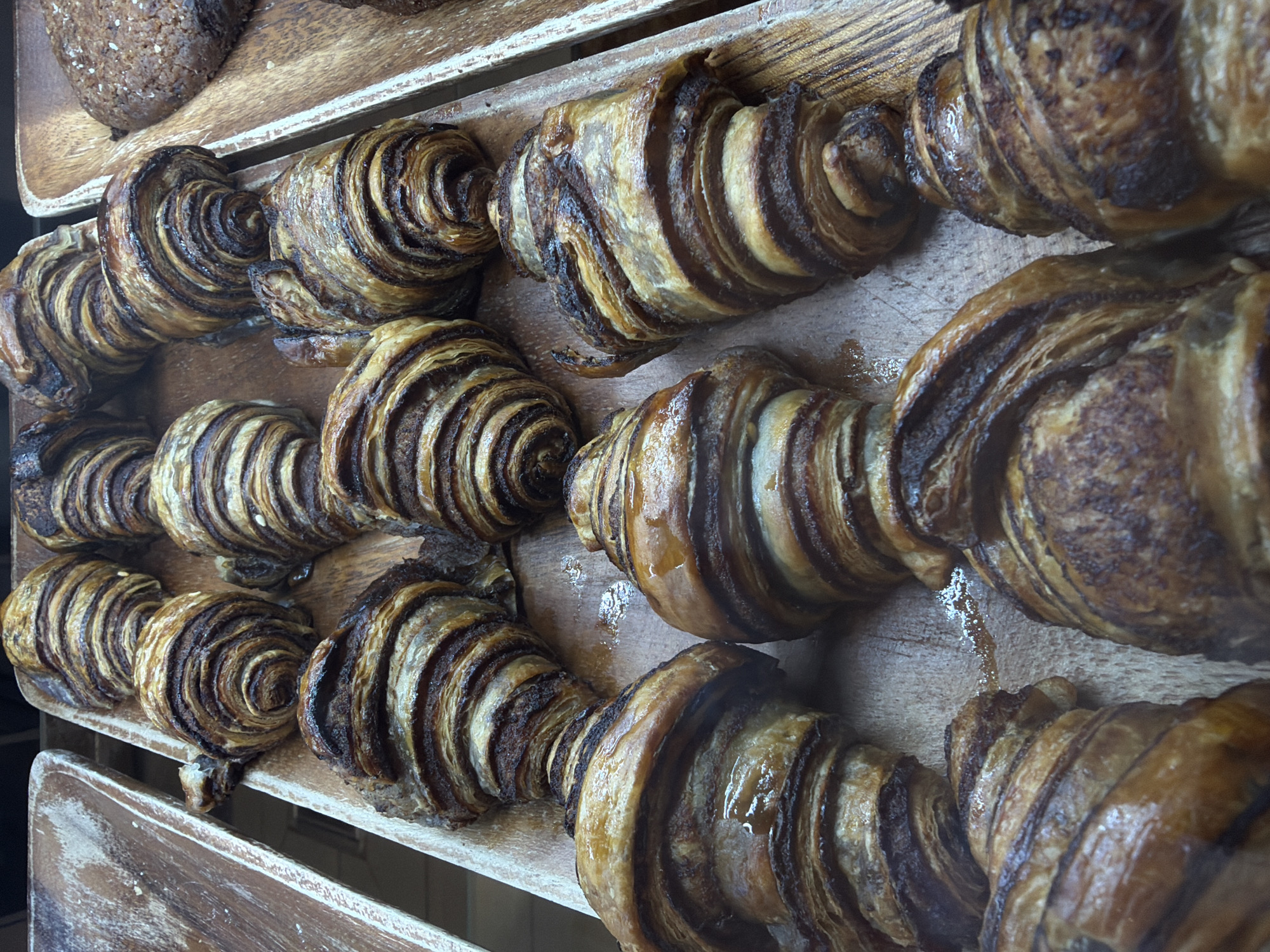 The image shows a wooden tray containing rows of chocolate babka pastries. The pastries are layered with chocolate filling, creating a swirling pattern when viewed from the side. The pastries appear to be displayed at a bakery in Tel-Aviv, Israel.