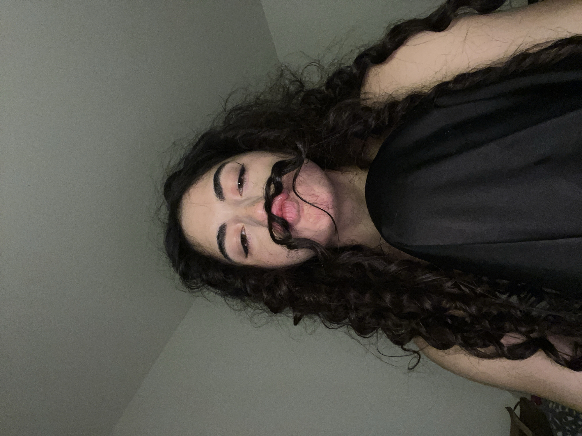 An image shows a young person with long, dark, curly hair and makeup. Their hair is styled into a mustache shape over their upper lip. The person is wearing a black garment. The background is a plain, lightly colored wall with a visible corner. The image appears to be taken indoors in a dimly lit room. The location is not specified as Chotěšov, Czechia.