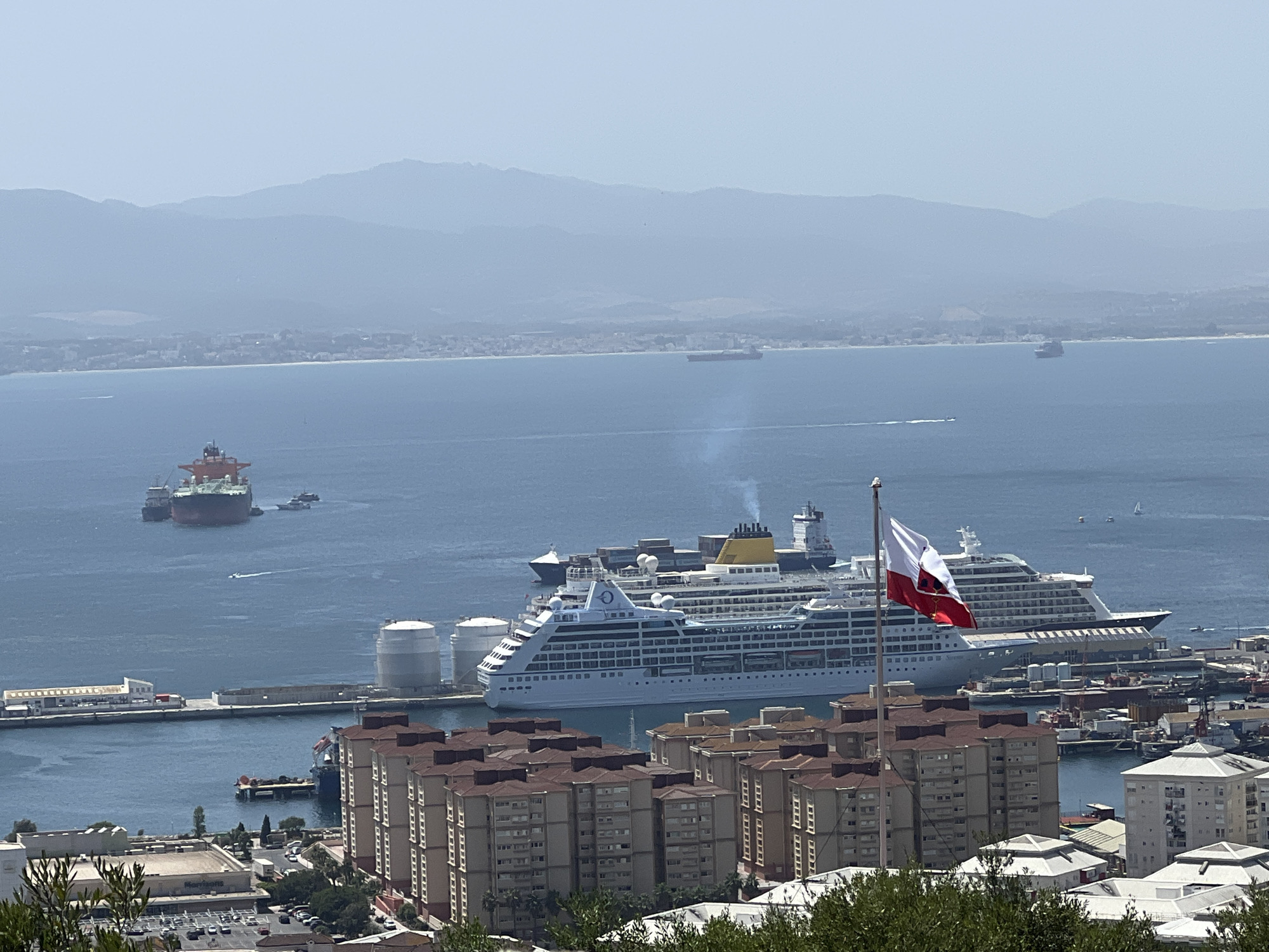 The image shows a wide view of the city of Gibraltar, Gibraltar. In the foreground, several apartment buildings with red tiled roofs are visible. A flag pole with the flag of Gibraltar is also in the foreground, with the flag waving. In the middle ground, a large cruise ship is docked in the harbor, with several other smaller boats and ships around it. In the background, there is a body of water, and in the distance, there are mountains. The sky is clear and blue. The overall scene is one of a bustling port city on a sunny day.
