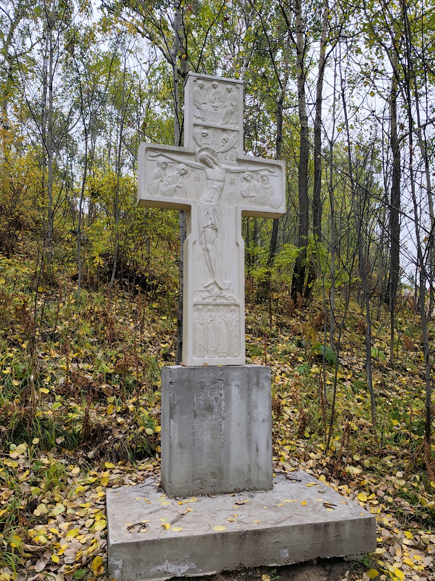 The image depicts a large, carved stone cross standing on a concrete pedestal in an outdoor setting. The cross features relief carvings of biblical scenes, including the crucifixion of Jesus Christ. The surrounding environment is a forest with deciduous trees, many of which have yellowing leaves, indicating an autumn season. The ground is covered with fallen leaves. The cross and pedestal appear to be weathered. The image was taken in the city of Nikola, Russia, likely in a park or a memorial site. There are no people or animals visible in the image. The lighting suggests an overcast day.