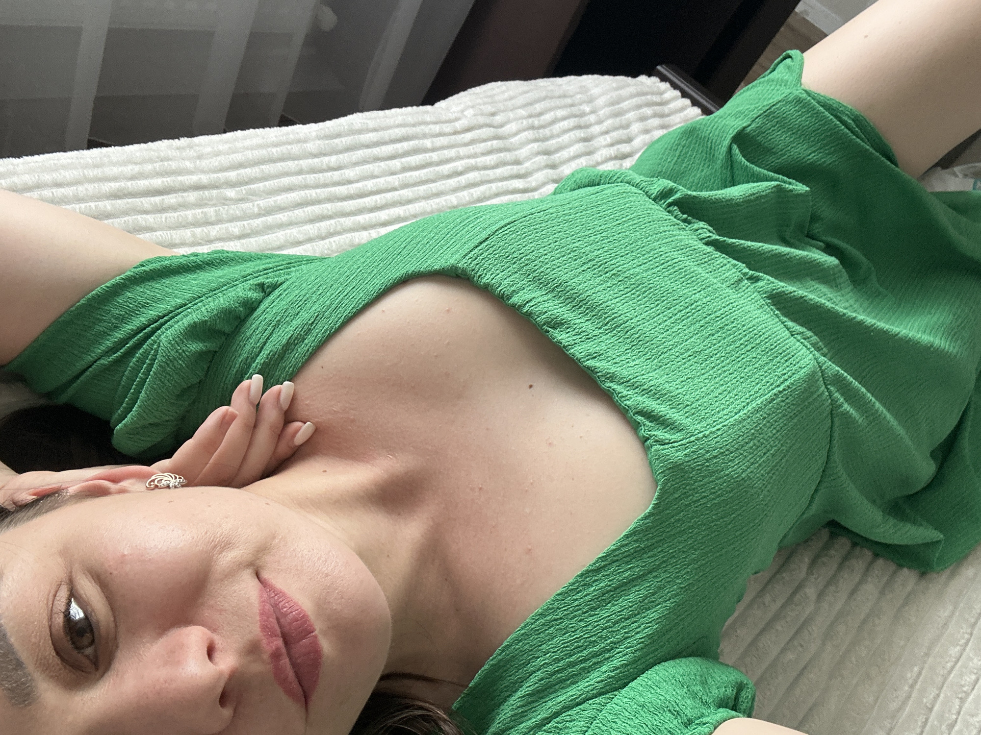 A woman lies on a white, textured bed in a green dress with a low-cut neckline. Her head is tilted toward the camera, her right hand resting near her forehead. A ring is visible on her hand. The photo appears to be taken indoors. There is no verifiable evidence to support the location as Chaikovsky, Russia.
