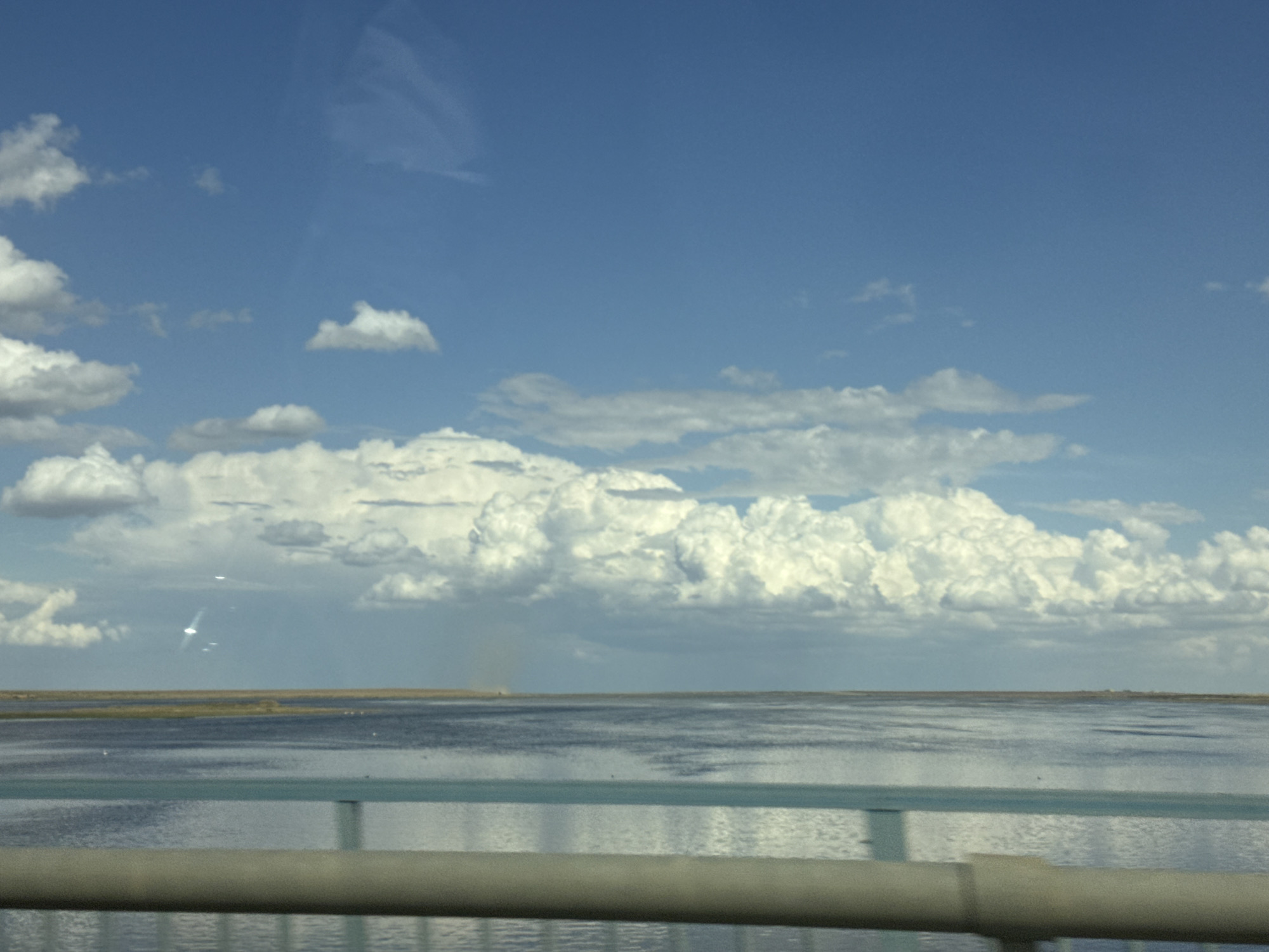 This is a landscape photograph taken from a moving vehicle, likely a car, as indicated by the presence of a railing in the foreground that appears to be part of a bridge or overpass. The scene depicts a wide expanse of calm, blue-grey water under a bright, partly cloudy sky. The horizon is flat, with a low, dark strip of land visible in the distance, suggesting a coastal or deltaic environment. Scattered white birds are visible on the water.
The sky is dominated by a vibrant blue, with fluffy white cumulus clouds in the middle ground and less defined, wispy clouds higher up. There are also some unusual bright, lens flare-like reflections in the sky, possibly caused by the sun or light sources outside the frame. The overall impression is of a clear and sunny day, likely during daylight hours. The water is smooth and reflects the sky and clouds, creating a serene and expansive feeling. The railing in the foreground, painted in muted blue and grey tones, creates a sense of depth and places the viewer within the scene. No people or specific man-made objects other than the railing are clearly identifiable. The environment suggests a natural, open setting.