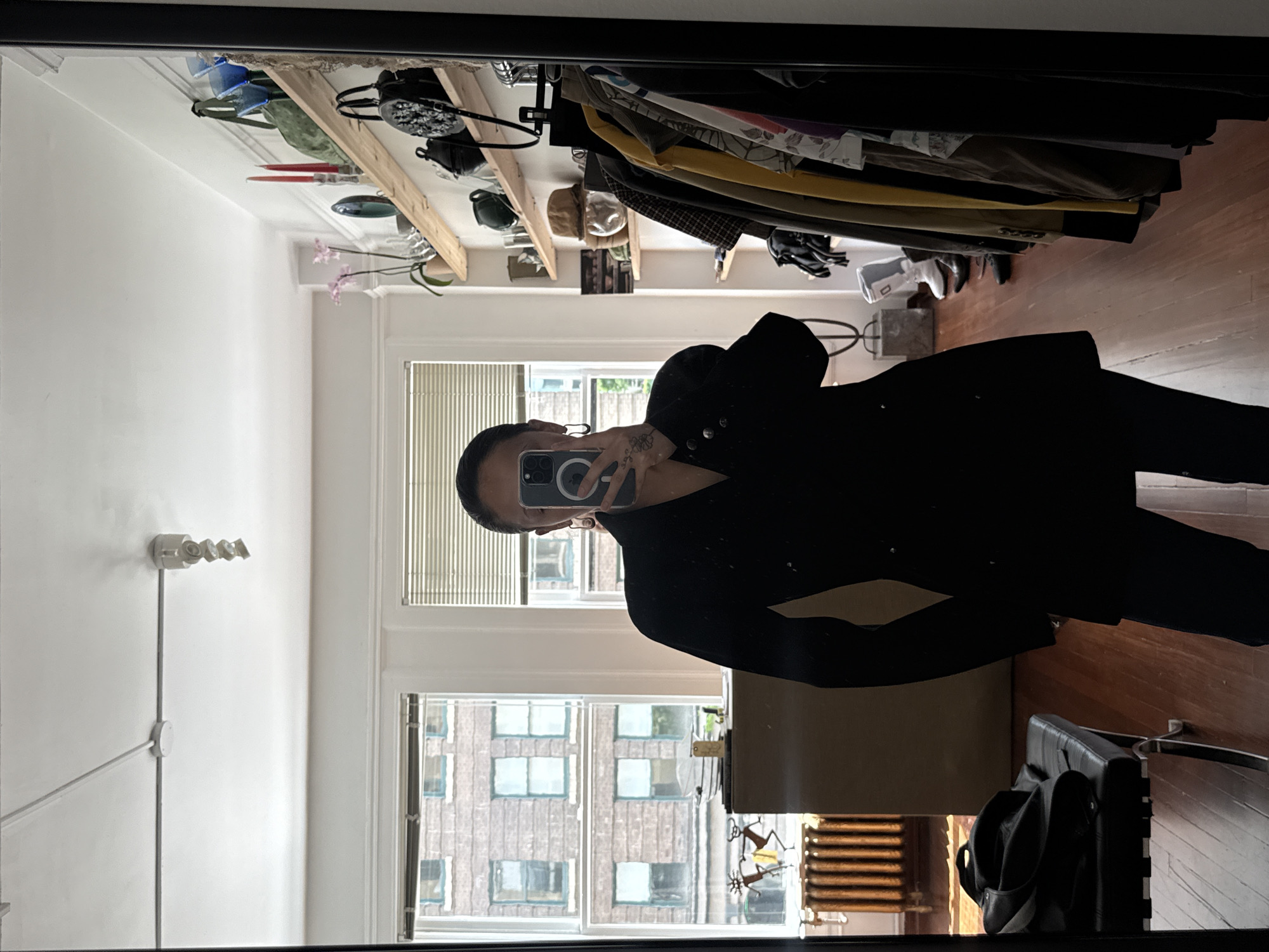 The image depicts a person taking a mirror selfie in what appears to be an apartment. The reflection shows a room with hardwood floors, a window facing what seems to be a brick building across the street, suggesting an urban setting. The window has a partial view of the building. A black leather jacket and pants are the main focus. The room also contains what appears to be a closet with clothes hanging inside, a wooden shelf with various items, and a light fixture on the wall. A black leather chair is also in the shot. There are no details that indicate if the photo was taken in Vancouver, Canada.