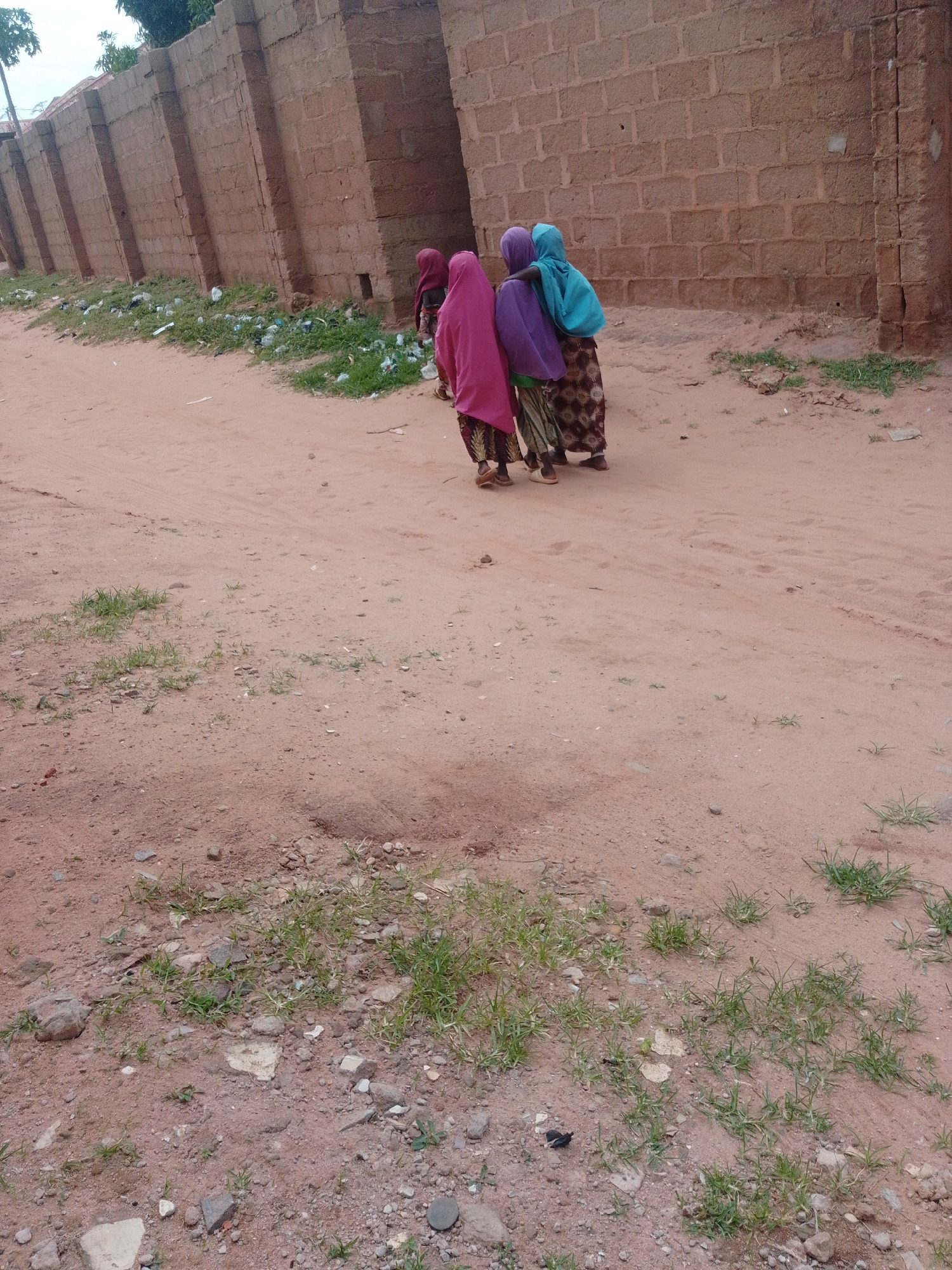 In the city of Unknown, Unknown, a group of three young girls stand by a dirt road next to a brick wall. The girls are wearing hijabs and long dresses. One girl is wearing a pink hijab, another a purple hijab, and the third a turquoise hijab. They are all looking away from the camera. The dirt road is sandy and there is some sparse vegetation growing along the side. The brick wall appears to be old and weathered. The scene is shot in daylight.