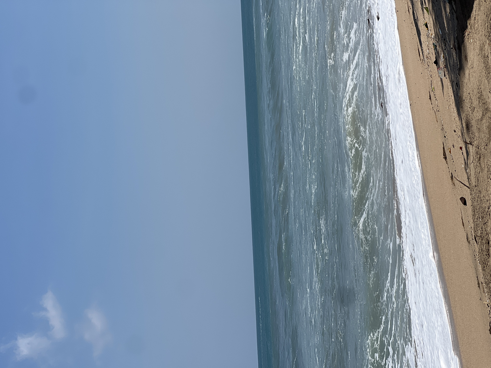The image shows a view of the ocean meeting a sandy beach. The sky is a clear blue with a few wispy clouds. The ocean is a deep blue-green, with white waves breaking on the shore. The sand is light brown and appears to be somewhat littered with small debris. The scene is a typical coastal landscape, likely depicting a beach in Kuta, Indonesia, given the context. There are no people or specific landmarks visible in the image. The overall impression is one of a natural, albeit somewhat unkempt, seaside environment.