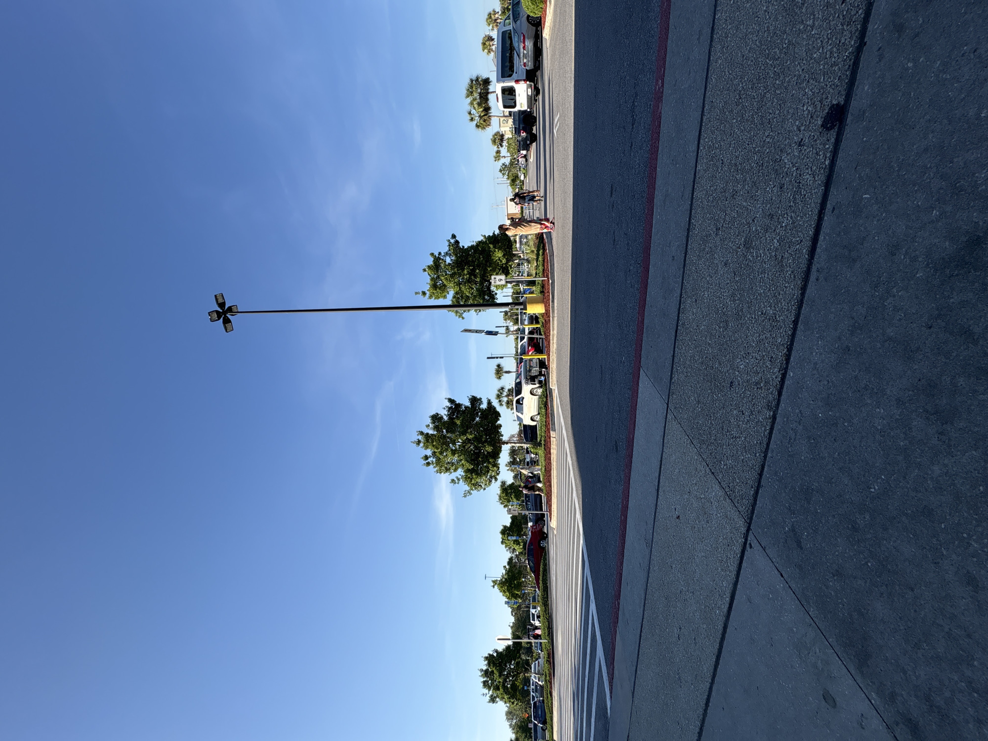 The image shows a sidewalk and a parking lot in Panama City Beach, Florida, United States of America. The sky is bright blue with a few wispy clouds. A tall street light pole stands on the left side of the frame. Trees line the parking lot, and cars are parked in the spaces. A red painted line separates the parking lot from the sidewalk. Two people are walking near the parking lot.