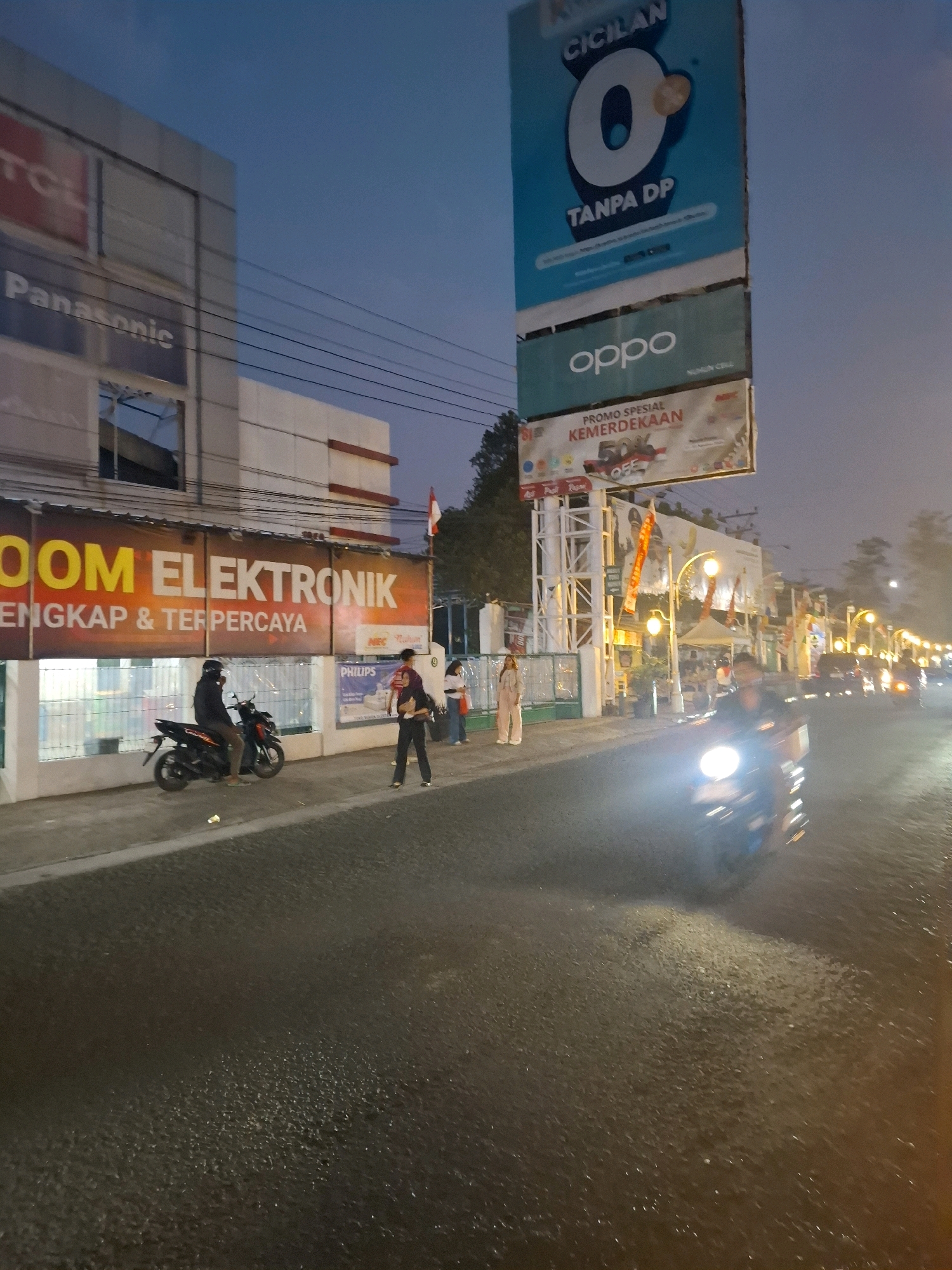 The image depicts a street scene at dusk in Sayang, Indonesia, in front of an electronics store. A large banner with orange and yellow lettering reads "ROOM ELEKTRONIK LENGKAP & TERPERCAYA". To the left, a building has signs for "TCL" and "Panasonic". A person wearing a black helmet and jacket is seated on a black motorcycle parked on the sidewalk. To the right of the motorcycle, three individuals are standing on the sidewalk. One person in a red shirt and black pants is facing away from the camera. Another person in a light-colored outfit is also facing away. A third person, also in light clothing, is standing beside them. Further down the street, the lights of other businesses are visible, and traffic is moving on the road. Several streetlights illuminate the area, and there are overhead wires spanning across the scene. A tall billboard structure is also present, displaying advertisements for "CICILAN O TANPA DP" and "OPPO". Another smaller banner reads "PROMO SPESIAL KEMERDEKAAN" with a 50% off graphic. The road surface appears wet, reflecting the lights.
