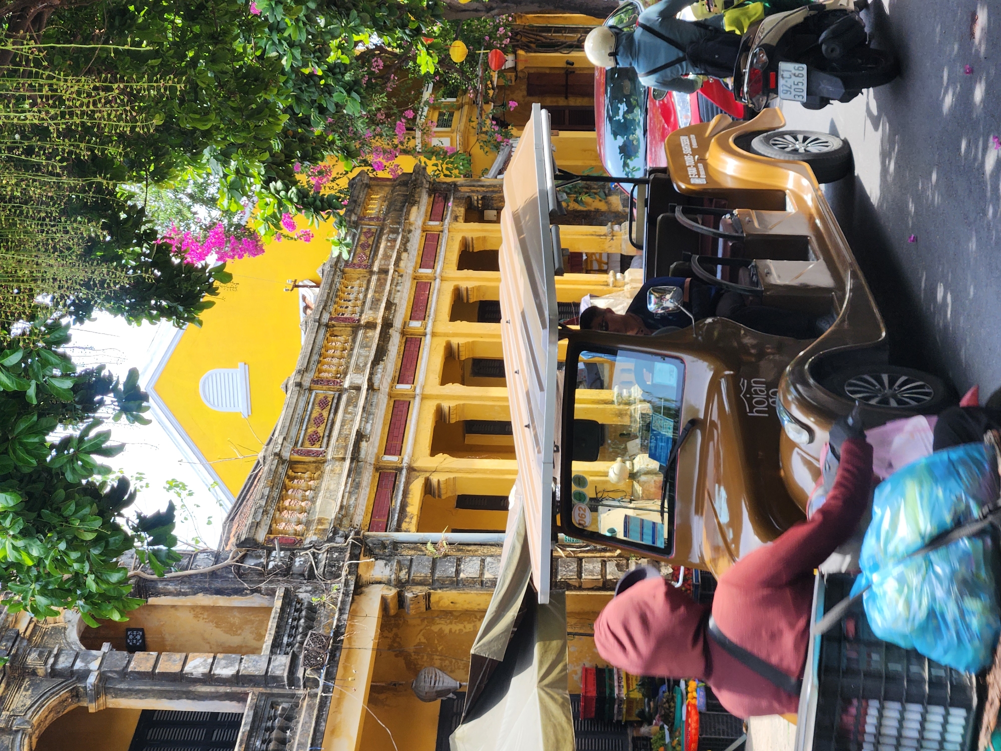 An exterior, eye-level, medium shot shows a street scene in Hội An, Vietnam, with buildings featuring aged yellow facades, ornate architectural details, and balconies adorned with classical balustrades. Lush green trees with delicate pink and dangling blossoms hang over the scene, casting dappled shadows on the asphalt. In the foreground, a woman wearing a hooded maroon jacket and black gloves is pushing a dark grey plastic crate filled with blue plastic bags. Behind her, a vintage-style, open-air electric cart with a tan roof is parked on the street. The cart displays the "HOIAN" logo on its side. Inside the cart, a person with short hair and sunglasses is seated in the driver's seat. To the right of the electric cart, a red car is visible. Further to the right, a person wearing a light blue jacket and a white helmet is riding a scooter. The scooter's license plate reads "92-C1 305.66". The overall atmosphere is that of a bustling marketplace or tourist area.