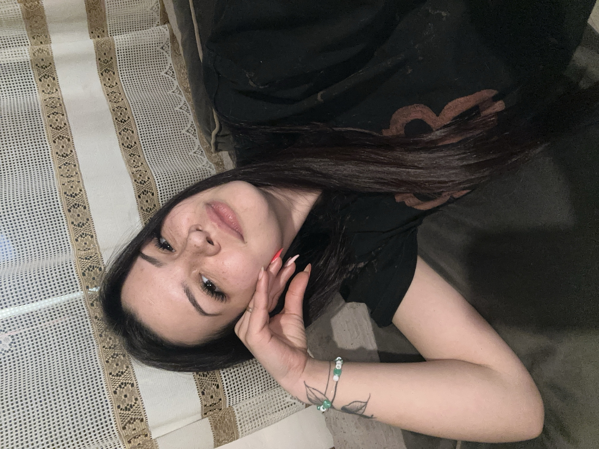 A person is lying on a couch or bed. They have long, dark hair, light skin, and are wearing a black t-shirt. They have one hand raised to their face, with red nail polish on their fingernails, and a beaded bracelet on their wrist. A drawing of a flower is also present on their arm. The background includes a crocheted blanket or tablecloth. The image does not contain any information suggesting it was taken in Chubra, Bulgaria.