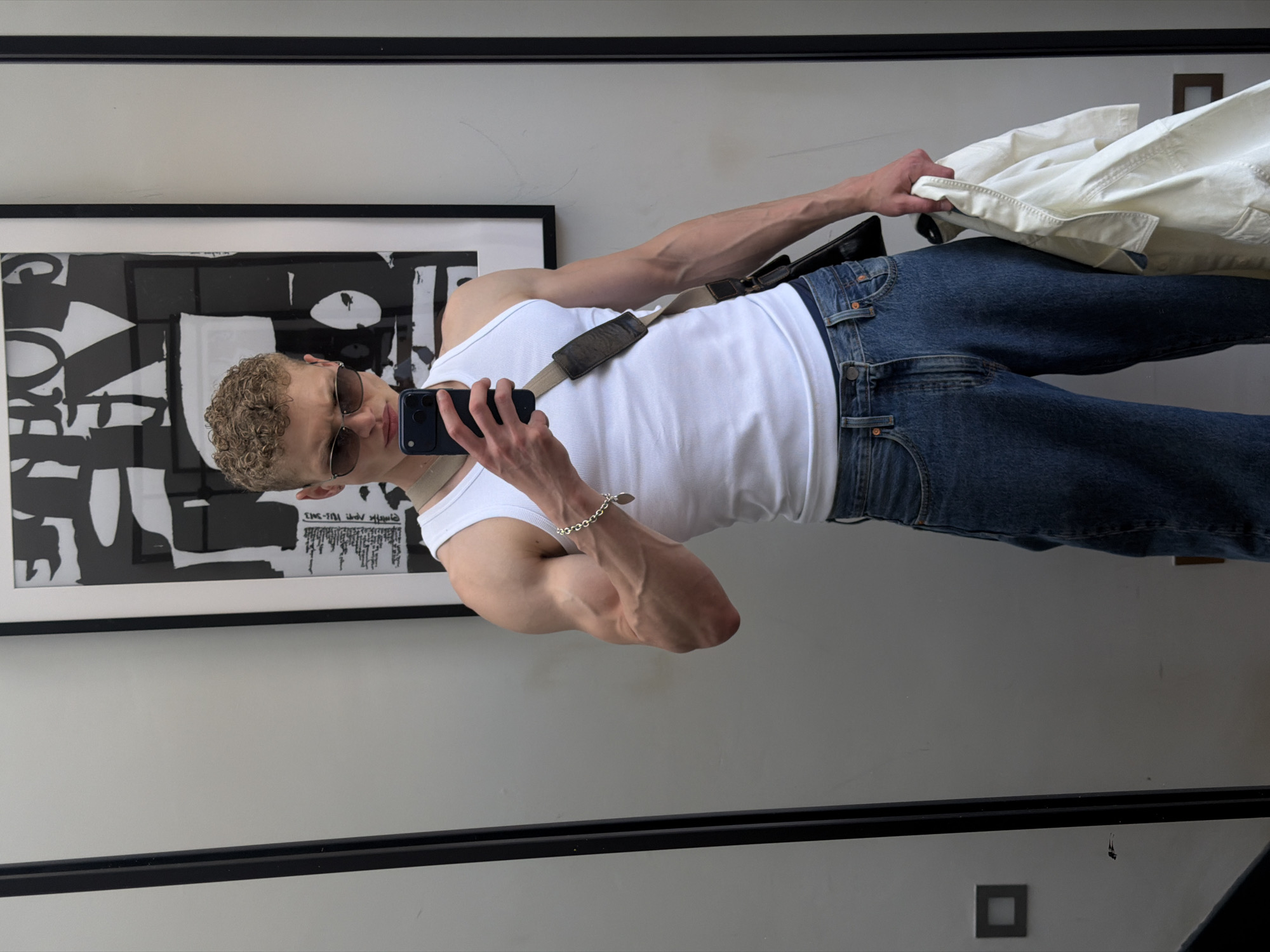 The image depicts a full-body mirror selfie of a young man, presented in a rotated, sideways orientation. The subject, who has short, curly light-colored hair, is wearing dark sunglasses, a plain white ribbed tank top, and dark blue jeans. A light-colored strap, possibly from a bag or camera, crosses his chest. He holds a dark-colored smartphone, likely an iPhone, in his right hand, angled towards his reflection to take the photo. His right arm, visible with prominent muscle definition and veins, sports a silver chain bracelet on the wrist. His left hand is seen holding a cream or white colored jacket, which is casually draped over his forearm.
The setting appears to be indoors, likely a modern apartment or a well-lit room in London, United Kingdom. Behind the man, a large framed piece of black and white abstract artwork hangs on a light grey wall. The artwork features bold, contrasting shapes and some stylized, partially legible text, including fragments like "ONE THS" and "YJ-3Y3". A black horizontal border, possibly part of the mirror frame or wall trim, is visible above and below the artwork. The overall lighting suggests it is daytime. The man's pose is confident and casual, typical of a fashion-oriented selfie.