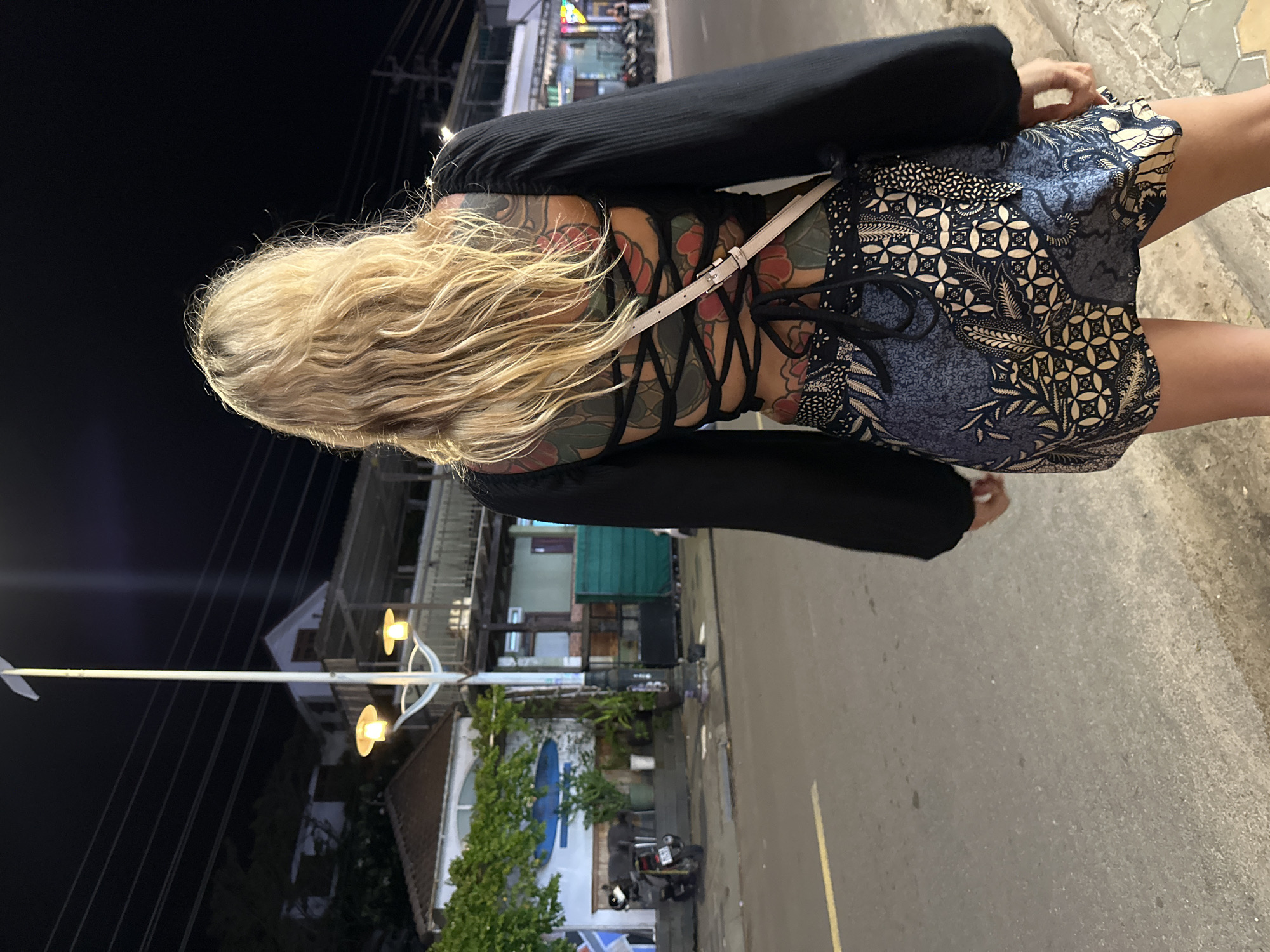 In Phu Thuy Ward, Vietnam, at night, a woman with blonde hair stands facing away from the camera, looking towards a street. She is wearing a black long-sleeved top with an open back featuring black lacing and a large floral tattoo on her back and lower torso. Her skirt is short and patterned with blue and white batik designs. She has a light-colored crossbody bag. In the background, a street light with two lamps illuminates the scene. Buildings with lights are visible behind the street light, and a motorcycle is parked on the left side of the street. The street is paved, with a yellow line marking the lane. The environment appears to be an urban area with a tropical feel, indicated by the vegetation.
