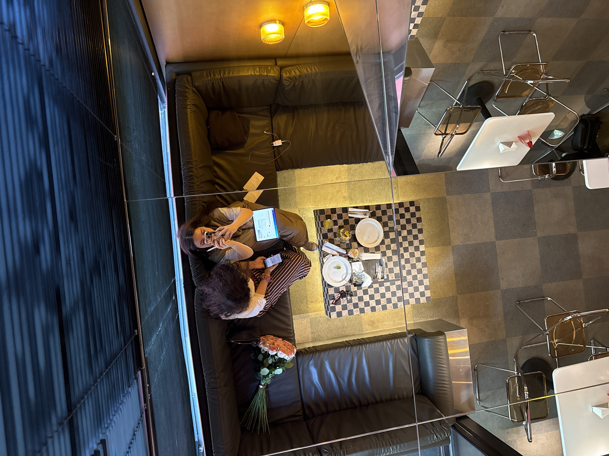 The image captures an overhead perspective of a space in Yerevan, Armenia. Two individuals are seated on a dark leather sofa, one using a phone and a tablet. A bouquet of roses is placed on the sofa near the other person. A table is set for a meal with dishes and cutlery. The flooring features a checkered pattern. The setting appears to be a restaurant with tables and chairs, partially seen with a reflective partition. The lighting includes overhead lights with visible reflection on the partition.
