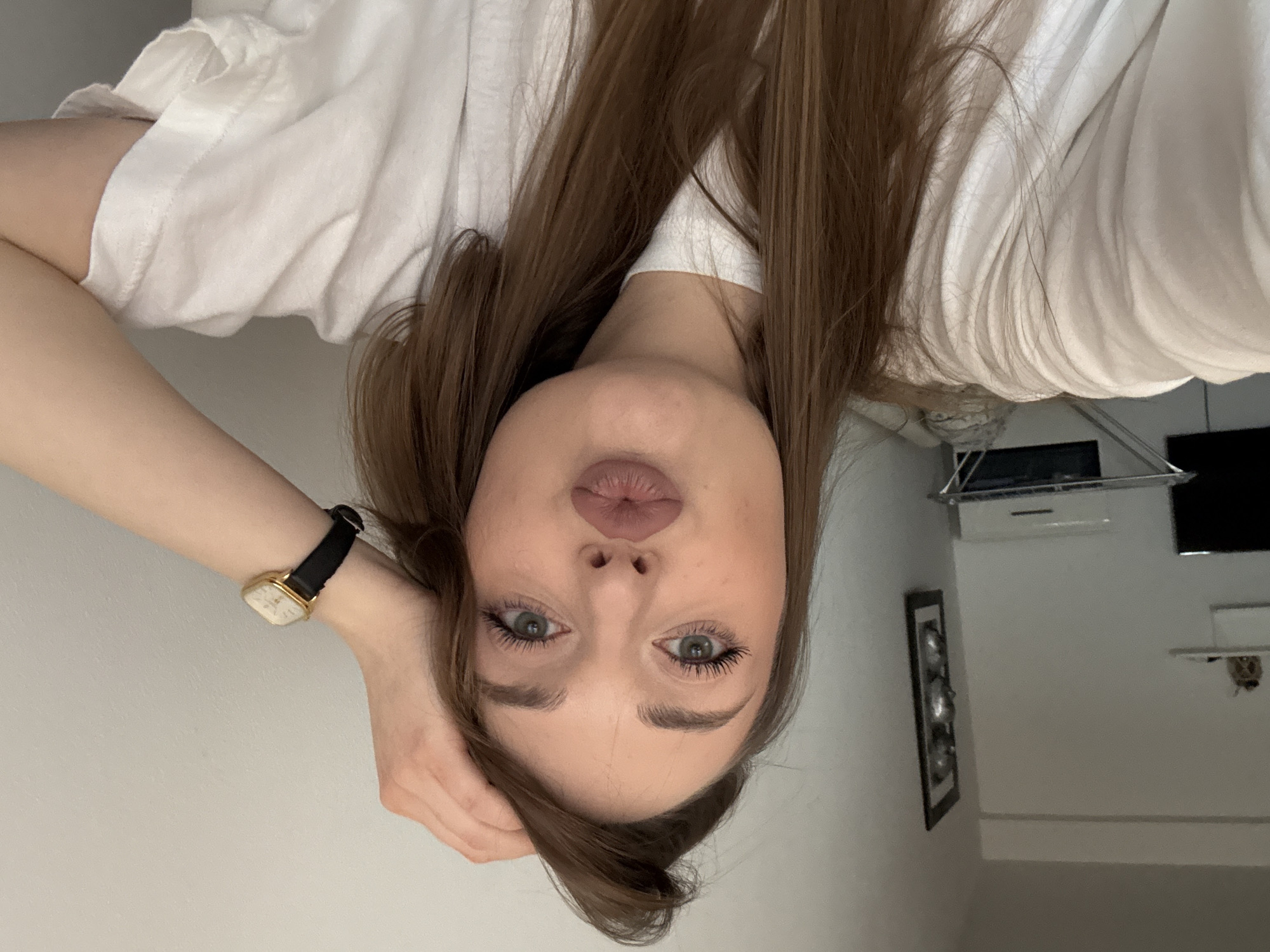 A young woman with long brown hair is posing for a selfie indoors. She is wearing a white shirt and a watch with a gold face and black strap. She has fair skin, light-colored eyes, and is puckering her lips slightly. She is resting her hand on her head. The background includes a white wall, a framed picture, a drying rack, and a television. There is no indication of the location in Kragujevac, Serbia.