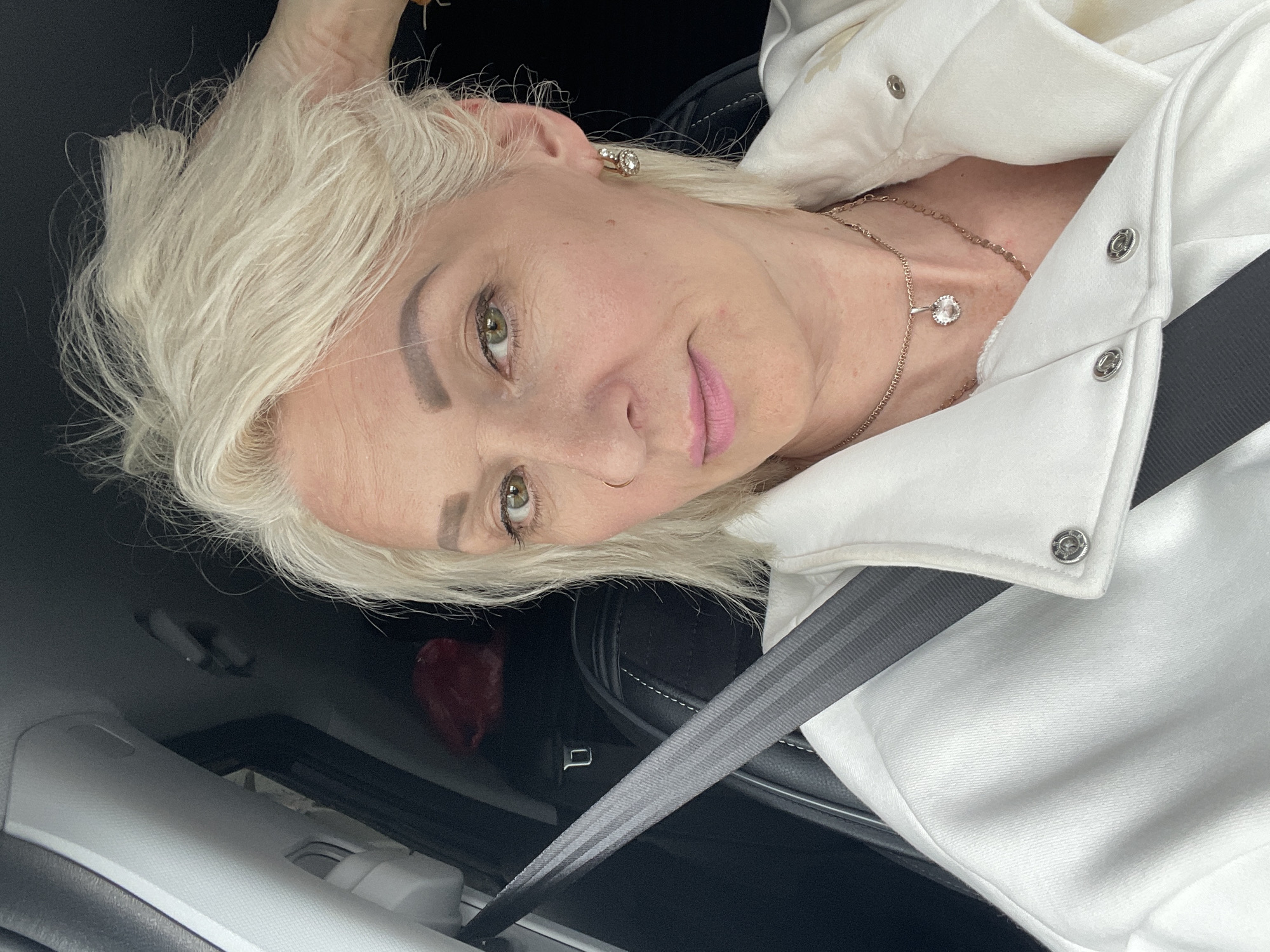 The image shows a close-up, selfie-style shot of a woman with blonde hair and light skin, wearing a white jacket and jewelry, seated in a car. Her right hand is touching her hair. The woman is wearing a necklace with a clear pendant and hoop earrings. A seatbelt is visible across her chest. The background is dark and indistinct, with a portion of a car seat visible. There is no information in the image to suggest a specific location in Gatchina, Russia.