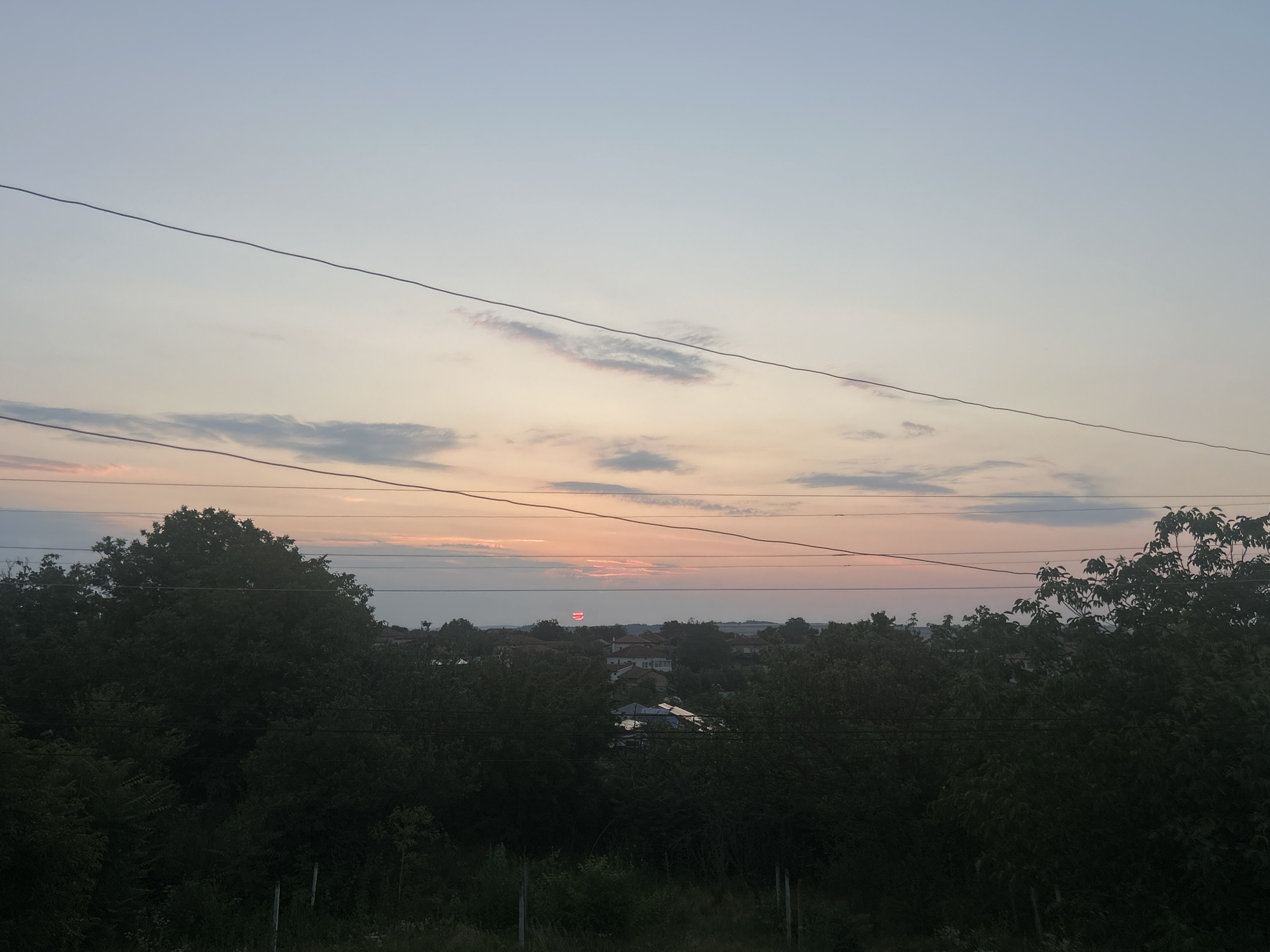 The image shows a sunset over the city of Loznitsa, Bulgaria. The sky is a gradient of pale blue to pink and orange, with thin clouds stretching across the horizon. In the foreground, dark green trees and foliage dominate the scene, with a few utility wires stretching across the frame. In the distance, a cluster of houses with red-tiled roofs are visible, partially obscured by trees. The sun, a small, red disc, is setting behind some of the houses and trees. The overall impression is one of a peaceful rural or semi-rural evening.