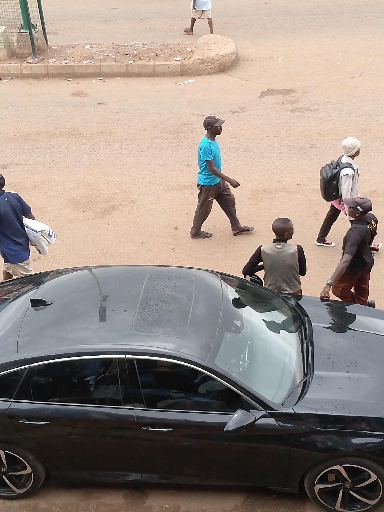 An overhead view captures a street scene in what appears to be a dry, possibly unpaved, urban or semi-urban environment in Bukuru, Nigeria. The foreground is dominated by the dark roof and side of a black sedan, possibly a Honda Accord, which has a sunroof and an antenna. Small water droplets or dust are visible on its surface.
Several individuals are present on the dirt road, walking in various directions. In the upper right, a person in light shorts and a white top walks away from the camera. Mid-left, a man wearing a blue t-shirt, brown pants, and a dark cap walks from right to left. Another individual with a light-colored top, dark pants, and a black backpack walks away from the camera in the mid-right, his hair appearing light or white.
Closer to the car, on the bottom left, a man in a blue shirt and light shorts carries a white bag with partially visible text, possibly "U" and another letter like "D". To the right of the car's hood, a person with short hair, wearing a dark long-sleeved shirt and a grey vest, faces away from the camera. Next to them, a man in a black short-sleeved top, brown pants, and a cap stands facing towards the car. The text "FZN" is clearly visible on his black shirt, suggesting a possible affiliation or brand. He wears a watch on his left wrist.
The ground is predominantly sandy or dusty brown, with a concrete curb separating the road from a slightly elevated area containing scattered debris and litter. A green metal fence or gate structure is partially visible in the upper left background. The scene is brightly lit by natural daylight, suggesting it is daytime. No distinct shadows are present, indicating midday or overcast conditions.