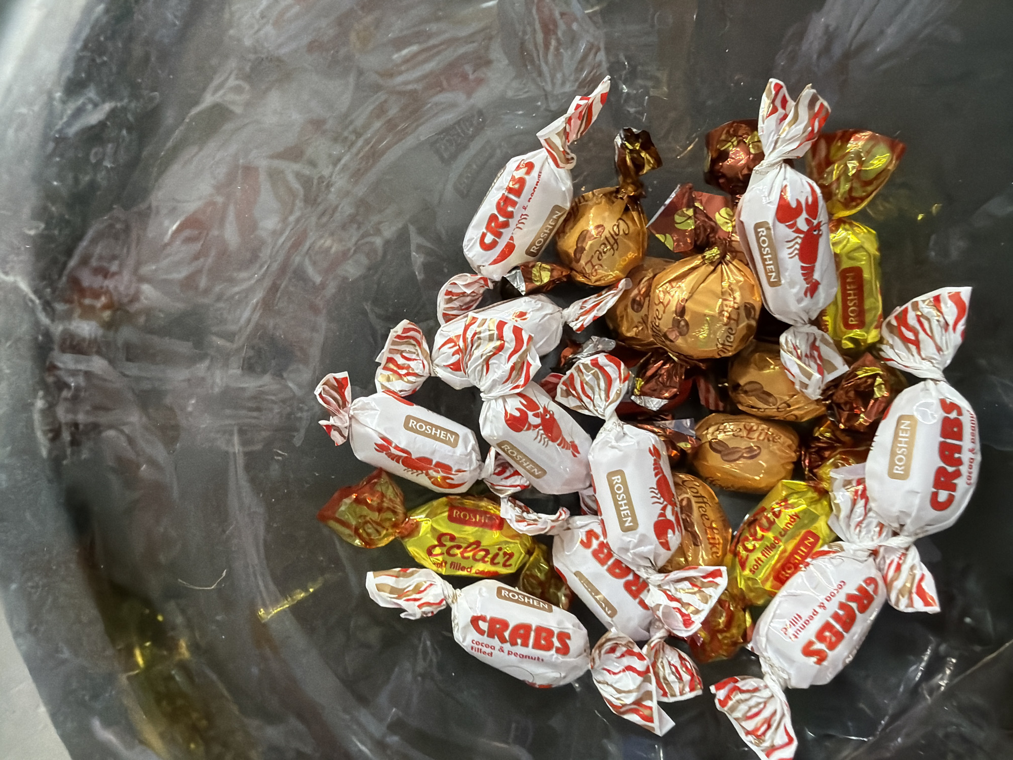 The image shows a pile of individually wrapped candies in a transparent plastic bag. The candies are a mix of brands and wrappers. Some wrappers are white with red and gold stripes and feature an illustration of a lobster and the brand name "CRABS" and "ROSHEN". Other wrappers are gold and brown with illustrations of coffee beans and the text "Coffee Like". There are also some yellow wrappers with red text that reads "Eclaire" and "ROSHEN". The candies are spread out within the bag, with some overlapping. The bag appears to be dark and slightly reflective, suggesting it is made of plastic. The image is taken from a top-down perspective. There are no people or identifiable locations visible in the image.