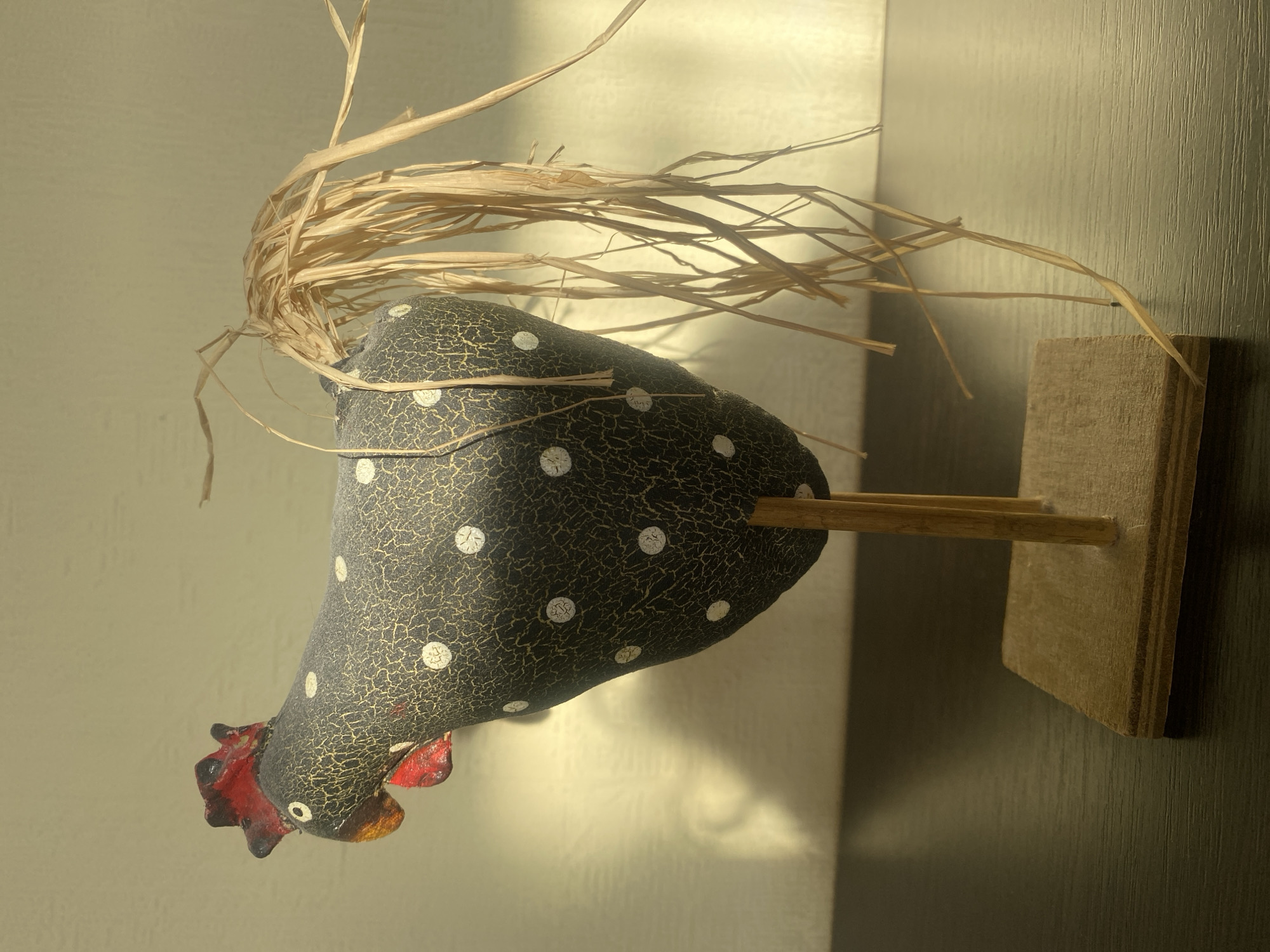A close-up shot of a decorative chicken figure with a black body, white polka dots, a red comb and wattle, and straw-like "feathers" on its head. It is mounted on a simple wooden stand with a dowel rod connecting the chicken to the stand. The background appears to be a wall or surface with subtle vertical texture and varying light conditions, suggesting indoor lighting or daylight entering from a nearby window. The composition emphasizes the texture and details of the chicken figurine. The location in Dnipro, Ukraine, is not apparent from the image.