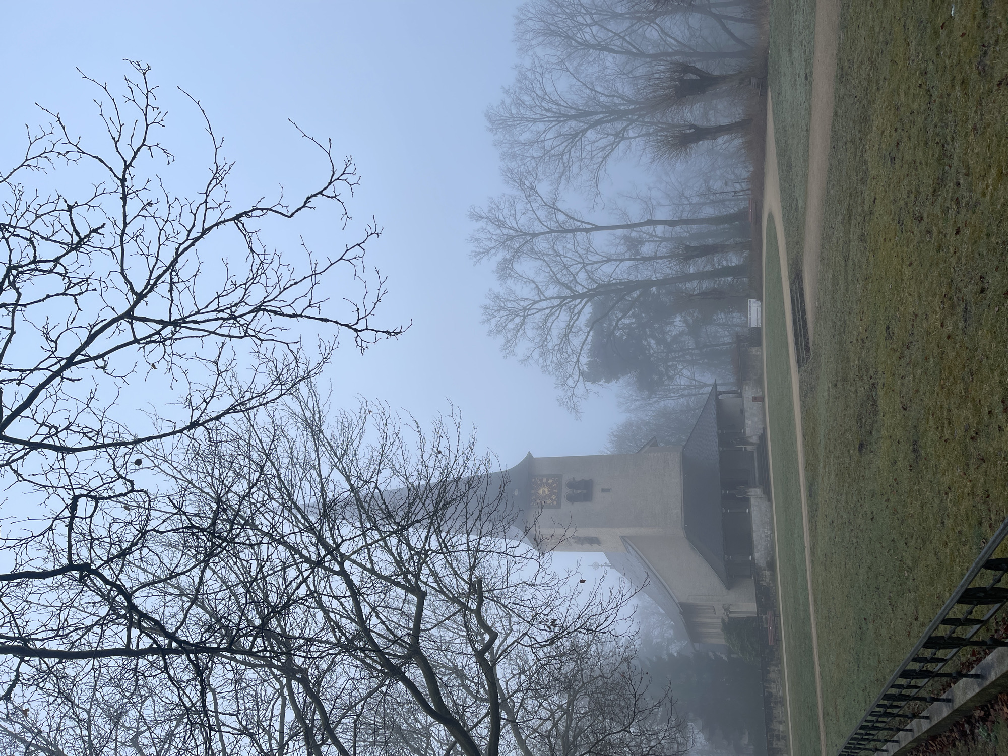 The image depicts a view of the Kaiser Wilhelm Memorial Church in Berlin, Germany, shrouded in fog. The iconic damaged spire is visible amidst bare trees. The surrounding park is grassy, and a pathway runs along the church. The fog obscures the background, giving the scene a muted, somber atmosphere. There are no visible people. A dark metallic fence is visible in the right side of the image. The clock on the church tower shows the time.