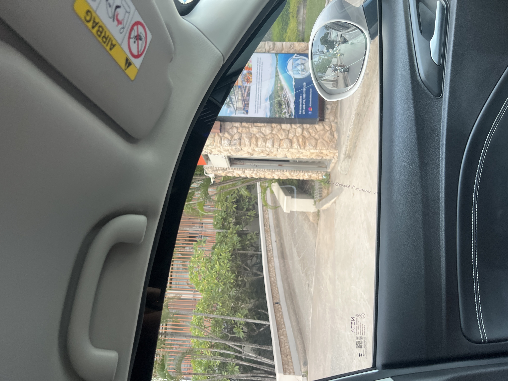 The image, taken from inside a vehicle, shows a view through the passenger side window and the side mirror. Outside, there is a stone-faced entrance with a large advertisement featuring a beach scene and contact information. A paved driveway leads away from the entrance, flanked by lush greenery and palm trees. The side mirror reflects a street scene with people on motorcycles. The interior of the car is visible, including a grab handle, a sun visor with an airbag warning label, and the car door panel.
