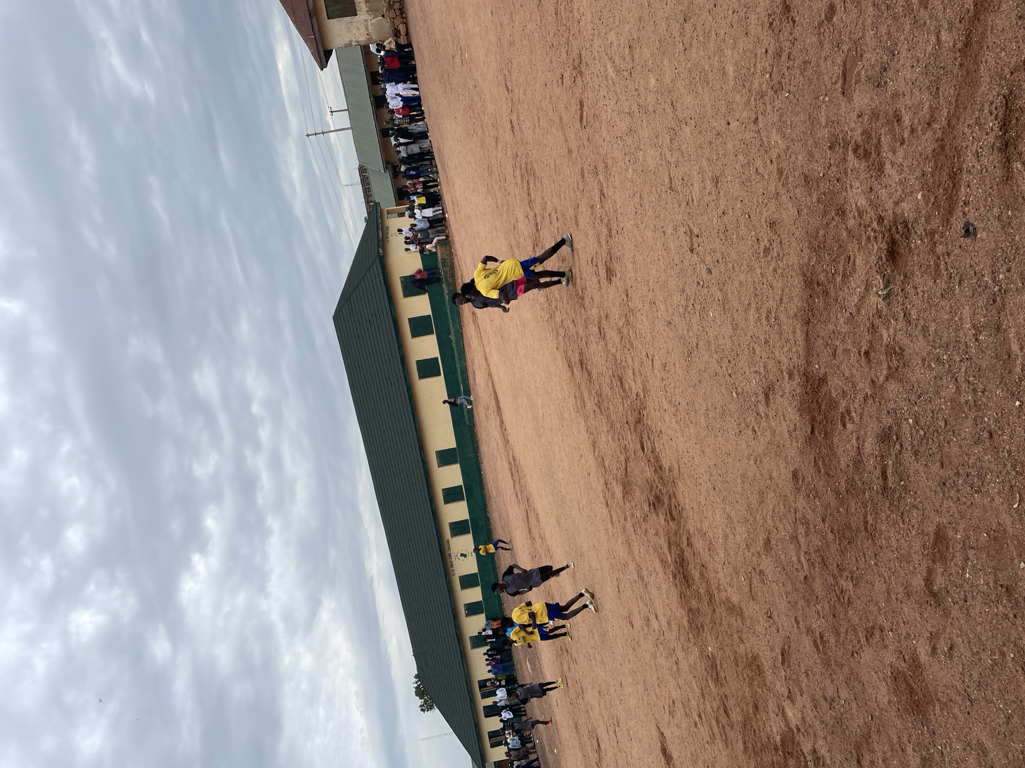 This image captures an outdoor scene, likely a schoolyard, in Jos, Nigeria. The foreground is dominated by a dusty, reddish-brown dirt field, indicative of the local terrain. A long, two-story building with a green-pitched roof runs along the left side of the frame. The building's walls are painted a pale yellow with dark green trim around the windows. Several dark green rectangular windows are visible. Text is visible on the building's facade: "E.C.N. PRIMARY SCHOOL, OJO-OLOWO."
A group of people are present on the dirt field. Some are standing and watching, while others appear to be engaged in an activity. In the middle of the field, two young men are interacting. One, wearing a yellow t-shirt and blue shorts, is being held from behind by another person. Further down the field, several other individuals in yellow and dark shirts are also visible, some appearing to be running or participating in some form of physical exercise. A larger group of spectators is gathered along the front of the building.
The sky above is overcast with a significant amount of cloud cover, suggesting a potentially dull or cloudy day. The lighting is diffused, and there are no strong shadows, which could indicate either early morning, late afternoon, or a generally overcast sky. The overall atmosphere appears to be one of community gathering and perhaps a school sporting event or physical education class.