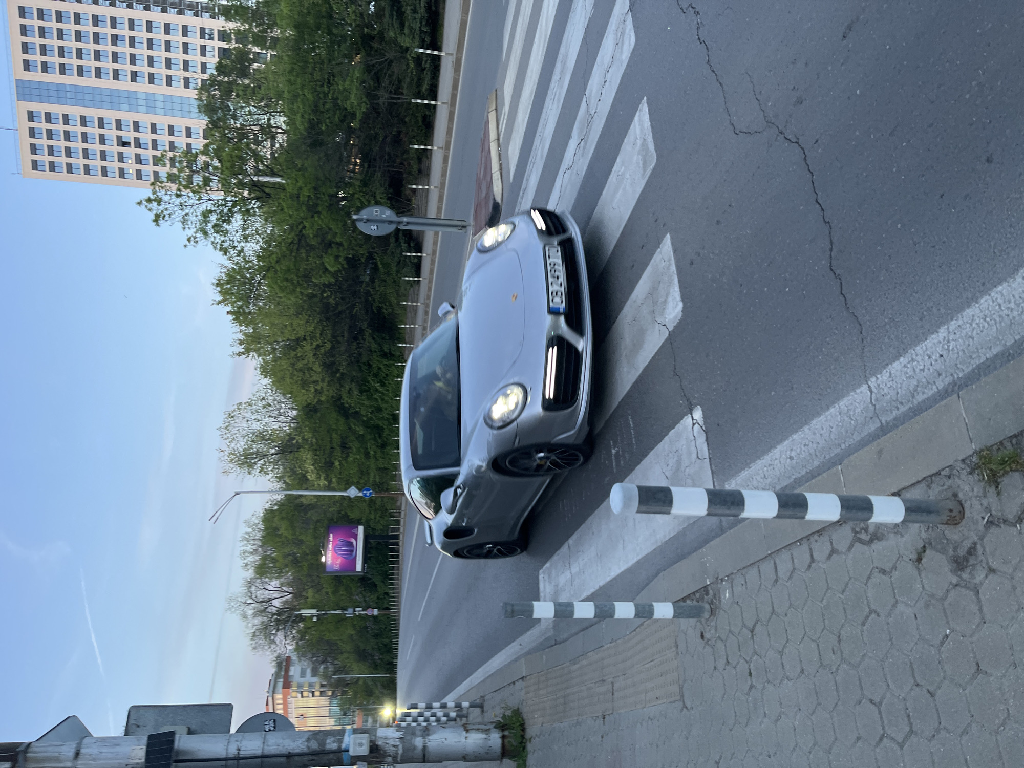 The image, rotated 90 degrees clockwise, captures an urban street scene during what appears to be late afternoon or early evening. A silver-grey Porsche Macan SUV is centrally positioned, either stopped or slowly moving on a white-striped pedestrian crosswalk. Its headlights are illuminated. The vehicle's license plate, "CB 2499 TC," is clearly visible, indicating registration in Sofia, Bulgaria.
In the background, a tall, modern, light-colored high-rise building with a grid of rectangular windows dominates the left side. A dense line of green trees runs along the road, partially obscuring the lower part of the building. The sky is a pale blue with faint contrails.
To the right, a grey sidewalk constructed from hexagonal pavers lines the street. Black and white striped bollards are placed along the sidewalk's edge near the crosswalk. The asphalt road shows visible cracks. Further down the street, a billboard with a purple hue is visible, featuring some indistinct white text that appears to say "create unique experiences." Various street signs and a lamppost are also present. The scene depicts a typical city environment with a mix of modern architecture and natural elements.