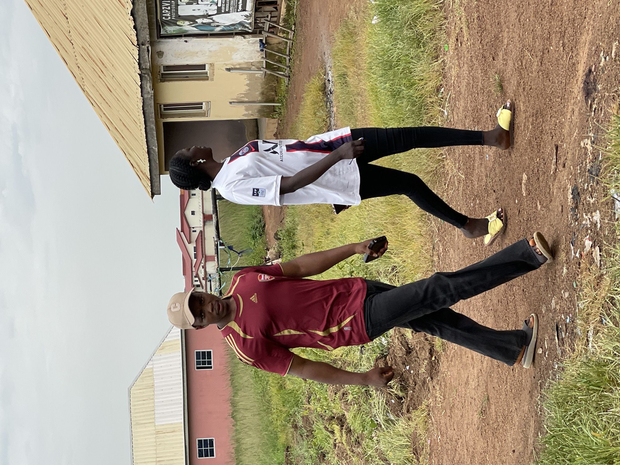 Two individuals are walking on a dirt path in Durumi, Nigeria. The person on the left is a male wearing a maroon and gold athletic shirt, dark pants, a beige baseball cap, and dark sandals. He is holding a dark-colored mobile phone in his right hand and looking towards the camera. The person on the right is a female wearing a white athletic shirt with blue and red accents, black leggings, and yellow sandals with decorative buckles. She is looking to her left. Behind them, there are several residential buildings with corrugated metal roofs and walls of various colors including pink and yellow. The ground is a mix of dirt, sparse green grass, and debris. The sky is overcast. A sign for "CAPZVIL PHOTOGRAPHY" is visible on the yellow building.