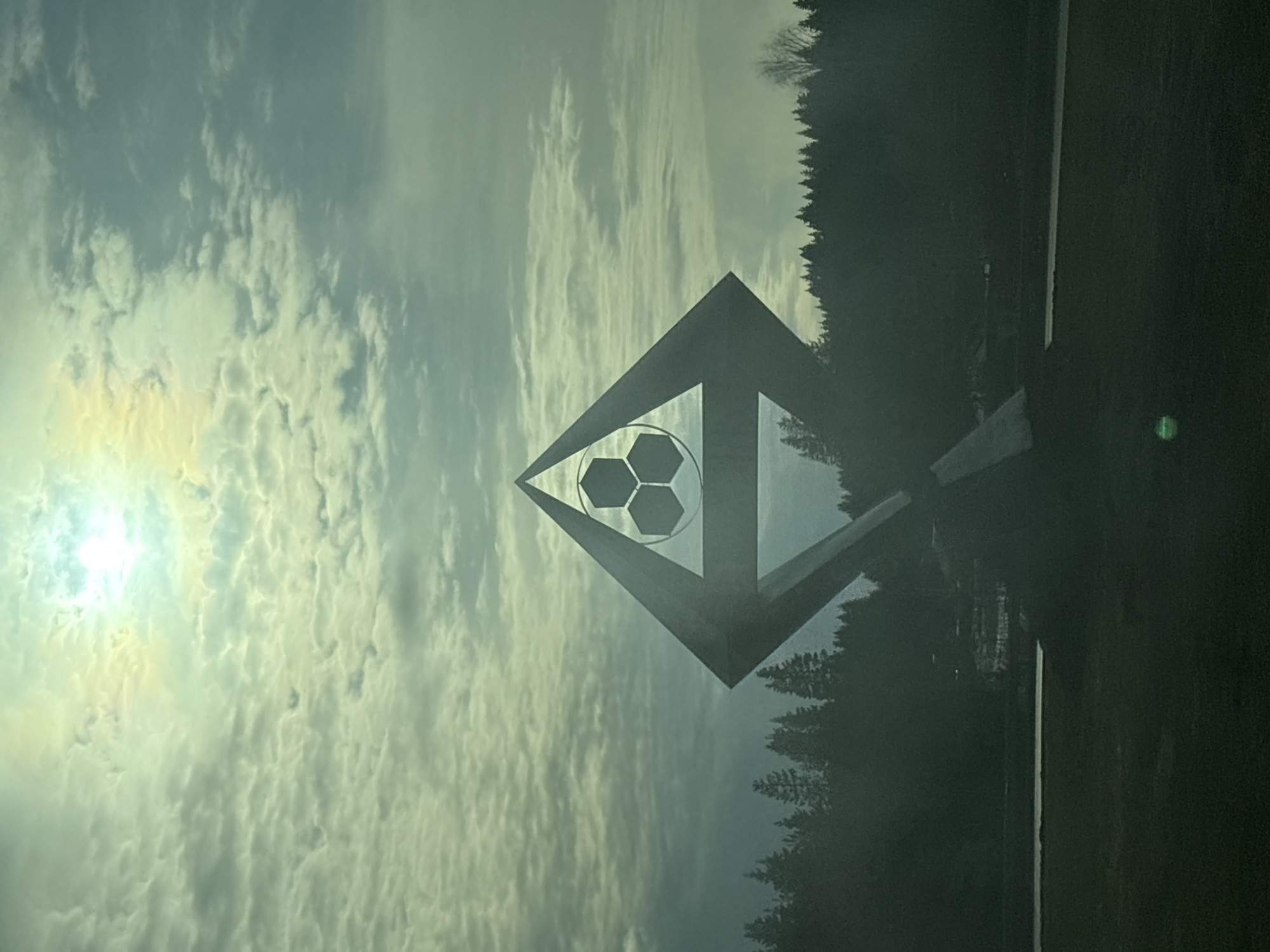 A sideways-oriented, slightly overcast daytime shot captures a soccer-themed monument against a backdrop of trees and sky. The monument features a diamond-shaped frame with a soccer ball design in the center. The sky is filled with scattered clouds and the sun peeks through them, creating a bright spot. The location is Berezniki, Russia.