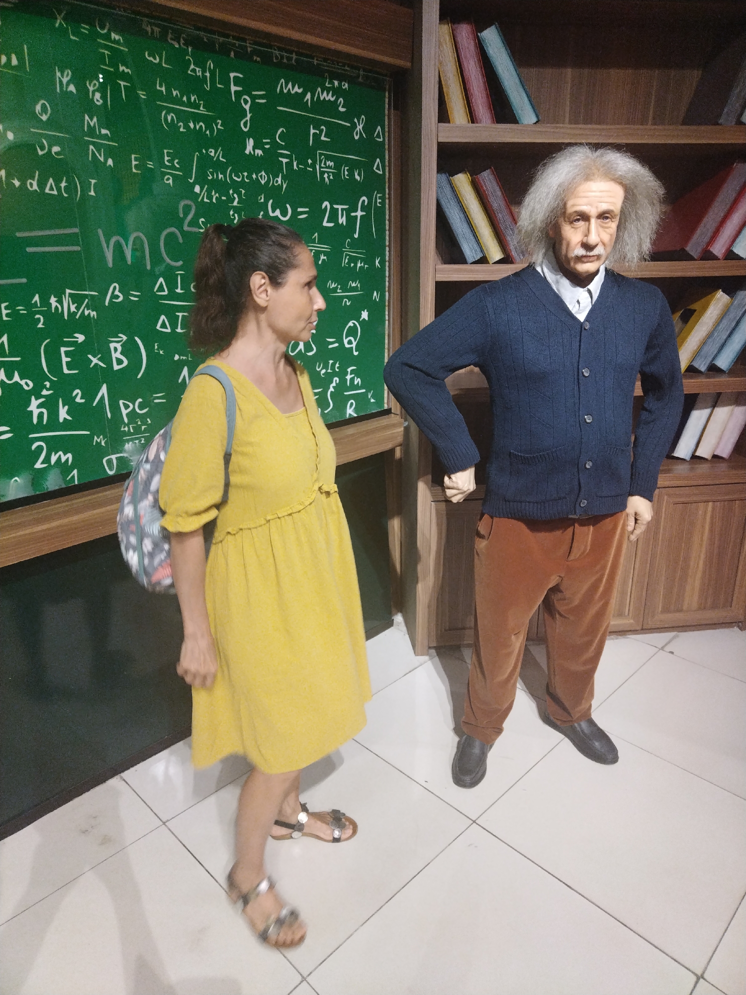 The image shows a woman posing next to an Albert Einstein wax figure. The woman is wearing a yellow dress and sandals and has a backpack over her shoulder. The Einstein figure is wearing a blue cardigan over a white shirt, with brown pants and black shoes. The figure is standing next to a bookshelf with decorative books, and behind the woman is a chalkboard covered in physics equations. The scene appears to be a wax museum or similar tourist attraction. The location is Unknown, Unknown.
