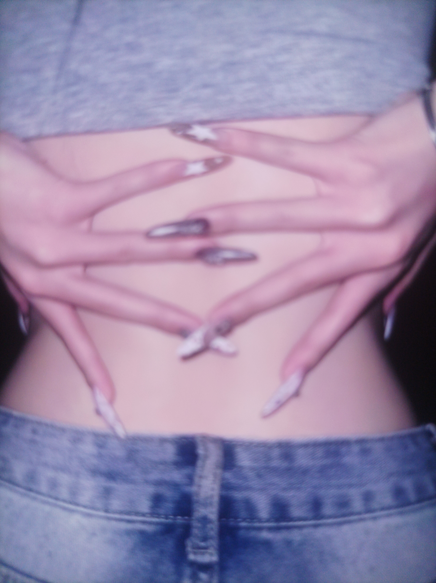 An extreme close-up shot focuses on the midsection and hands of a person wearing a grey crop top and blue jeans. The person's hands are intertwined across their lower back, with long, pointed fingernails decorated with a star pattern. The lighting is soft, with a pinkish hue, and the image is slightly out of focus, creating a dreamlike or ethereal effect. The background is dark and indistinct. The scene is devoid of any contextual clues about its specific location within Unknown, Unknown.