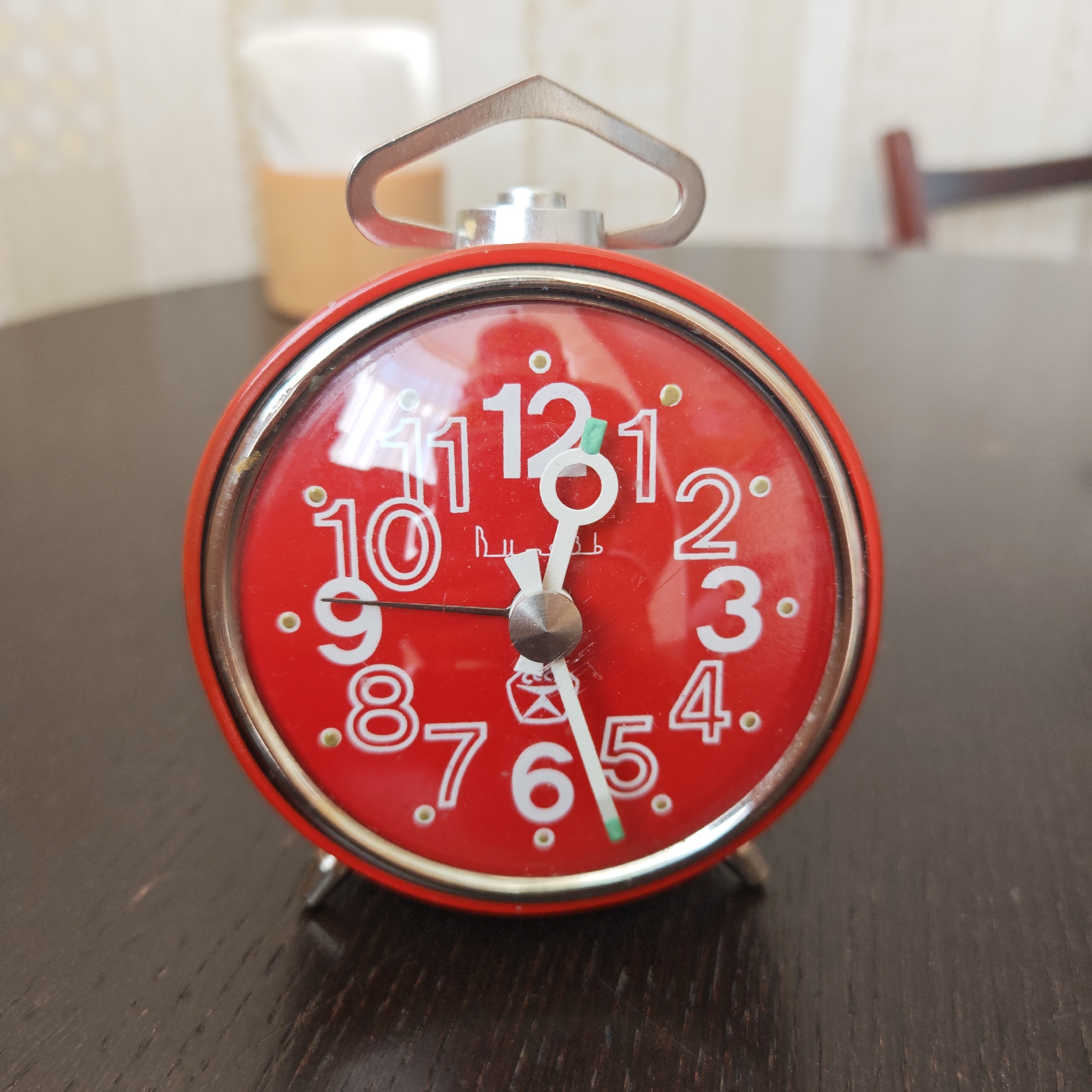 A red alarm clock sits on a dark brown, wooden surface. The clock face is red with white numbers. The minute hand is on the 6, and the hour hand is just before the 12. The alarm clock has a silver handle on top. The clock is sitting indoors. It is unknown if the location of the image is in the city of Taganrog City District, Russia.