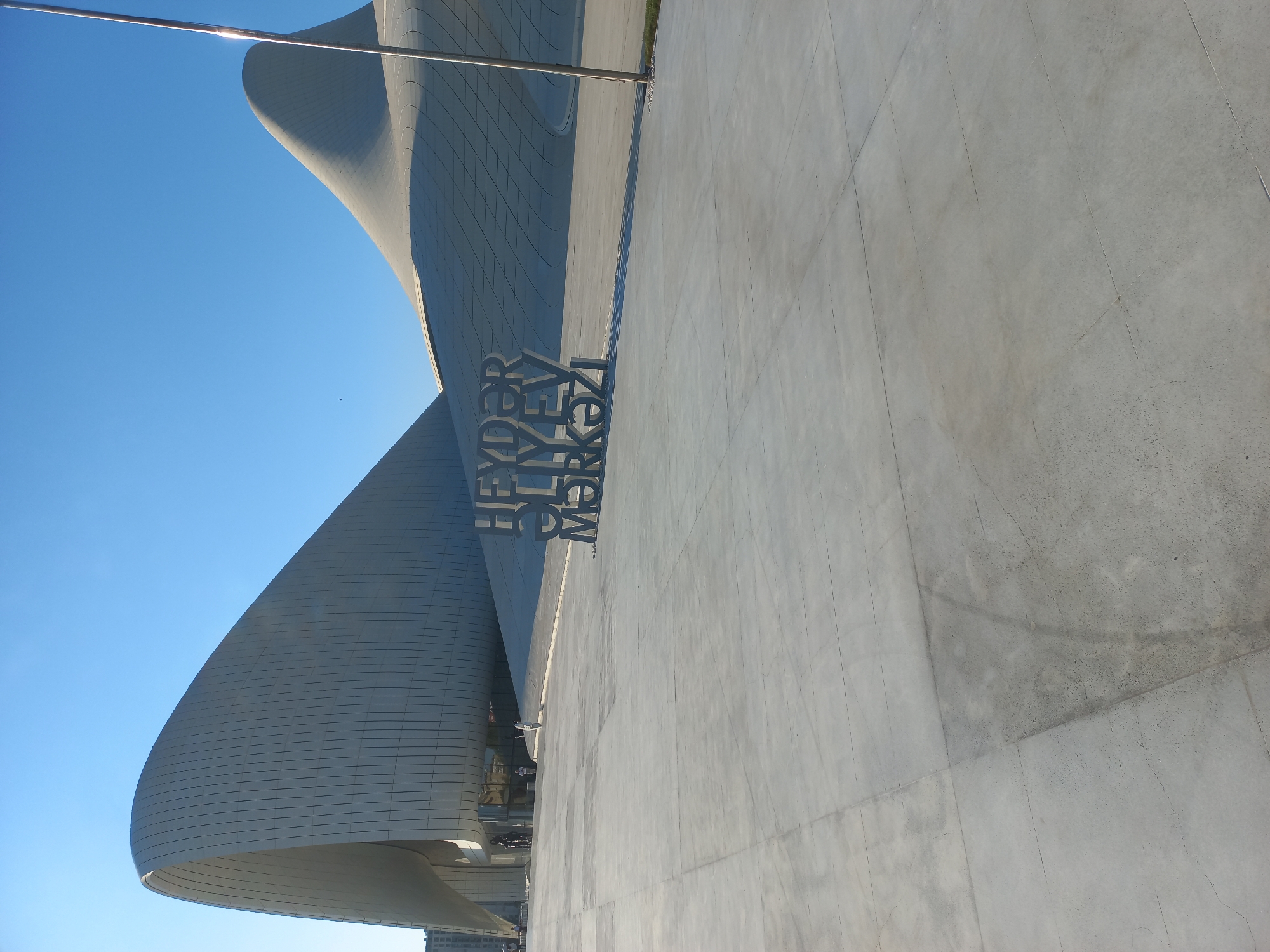 The image captures the Heydar Aliyev Center in Baku, Azerbaijan, an iconic architectural landmark known for its fluid and organic design. The building, constructed from white curved panels, dominates the background under a clear blue sky. In the foreground, large concrete slabs form a spacious plaza. A prominent metal sculpture spelling out "HEYDAR ALIYEV CENTER" in Latin script is visible on the plaza. A thin metal pole stands to the right of the sculpture, casting a shadow. A few individuals are present in the distance, near the entrance of the building, one on a bicycle. The scene suggests a sunny day, with clear lighting and minimal shadows on the plaza floor.
