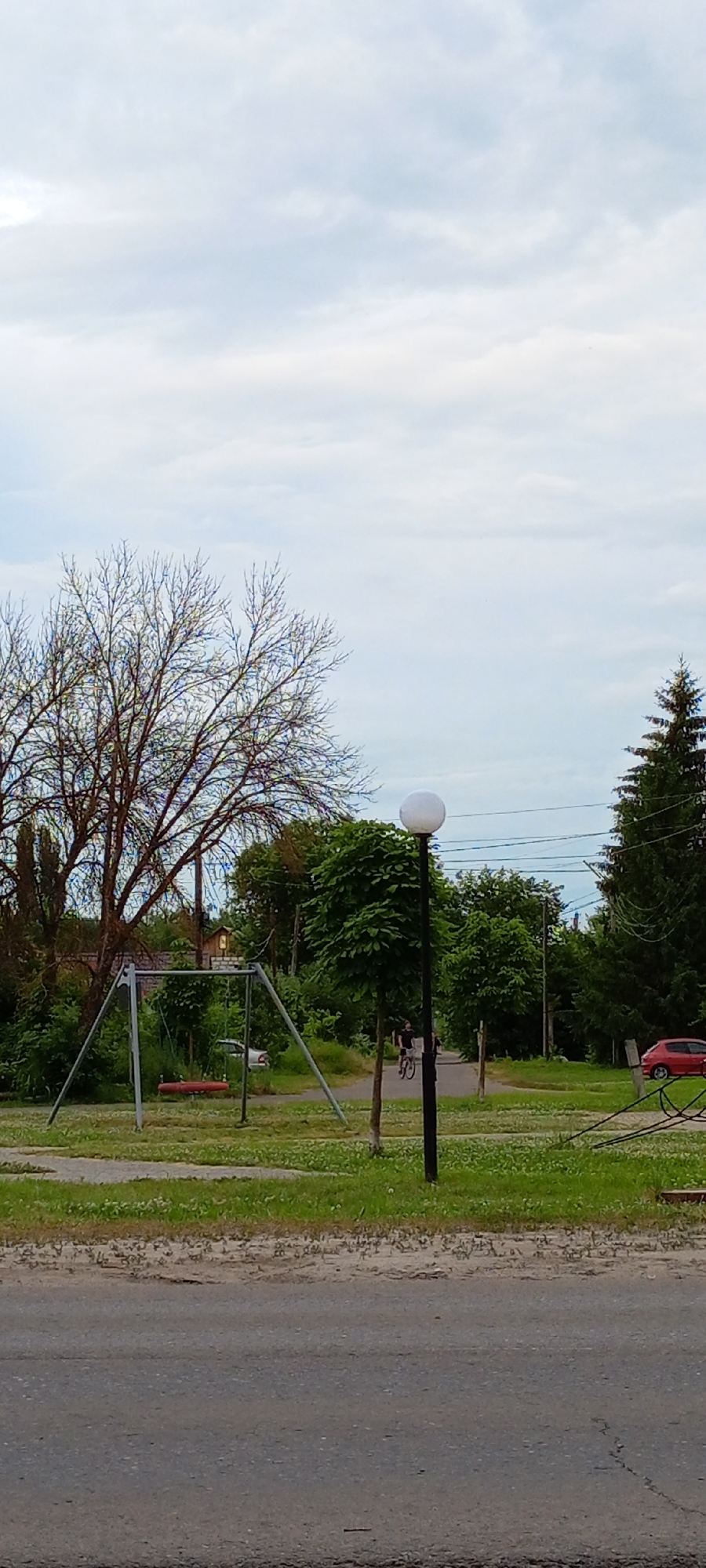 The image depicts a park scene in Kirsanov, Russia, under a cloudy sky. In the foreground, a paved road transitions to a grassy area. A swing set with a red seat is prominent on the left, situated near a bare-branched tree. A street lamp with a spherical white light fixture stands to the right of the swing set. Further back, a person is cycling on a path, and a red car is parked on the right side of the frame. Lush green trees and foliage surround the area, with power lines visible in the background.