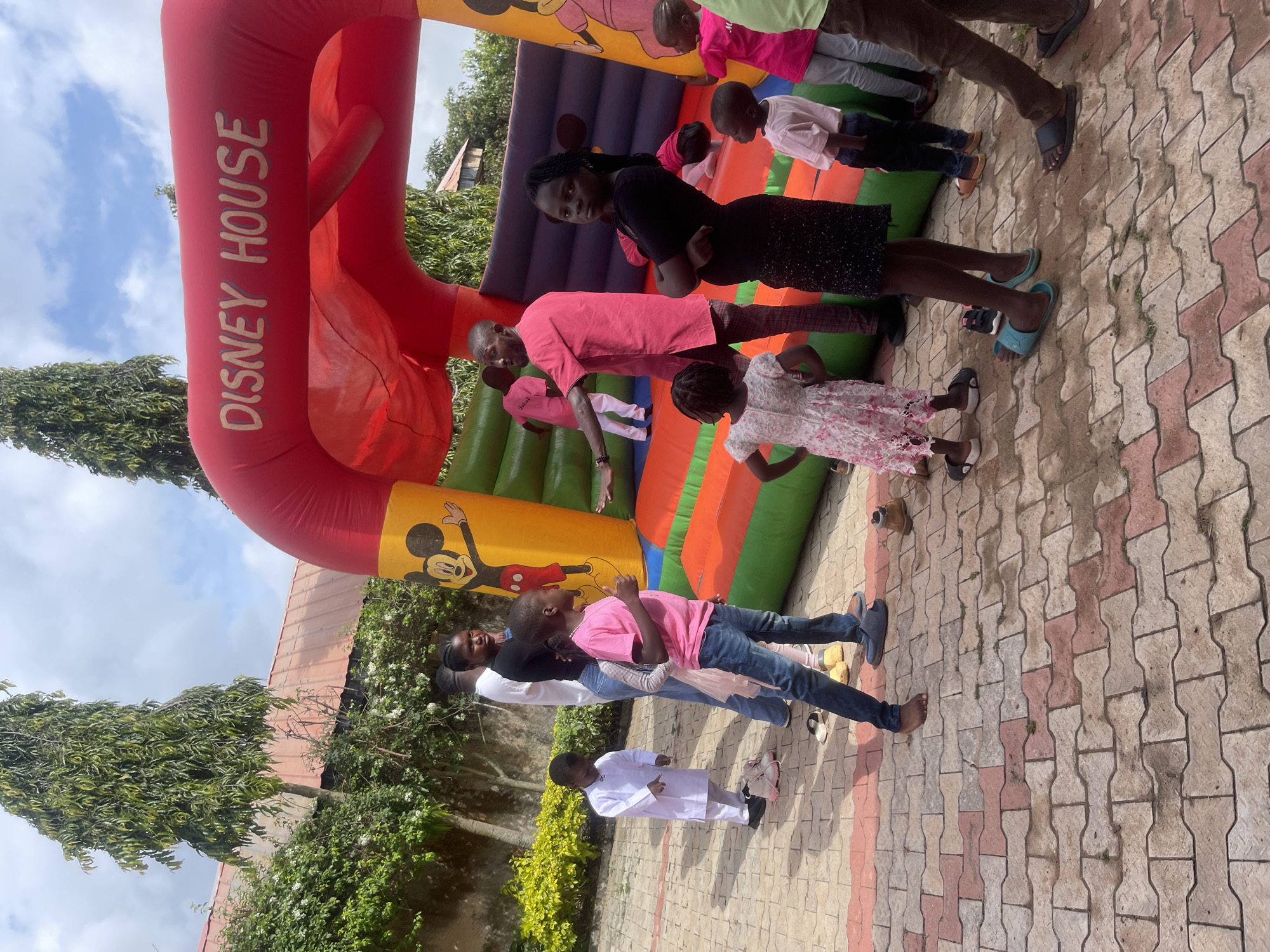 In Gura Top, Nigeria, a group of children and adults are gathered around a large red and yellow inflatable bounce house labeled "DISNEY HOUSE." The bounce house features images of Mickey Mouse. The ground is paved with interlocking bricks.
Several children, primarily young boys and girls, are visible, some standing near the entrance of the bounce house, others looking towards it. One boy in a pink shirt and blue jeans is giving a thumbs-up. A young girl in a white lace dress is looking away from the camera. Another girl in a black top and sparkly skirt is looking directly at the camera with her arms crossed. A man in a pink shirt and patterned pants is gesturing towards the bounce house, possibly addressing the children. Adults are also present, some observing the children and the bounce house. The environment appears to be outdoors, with trees and foliage in the background, and a partially visible building with a red roof. The sky is partly cloudy.