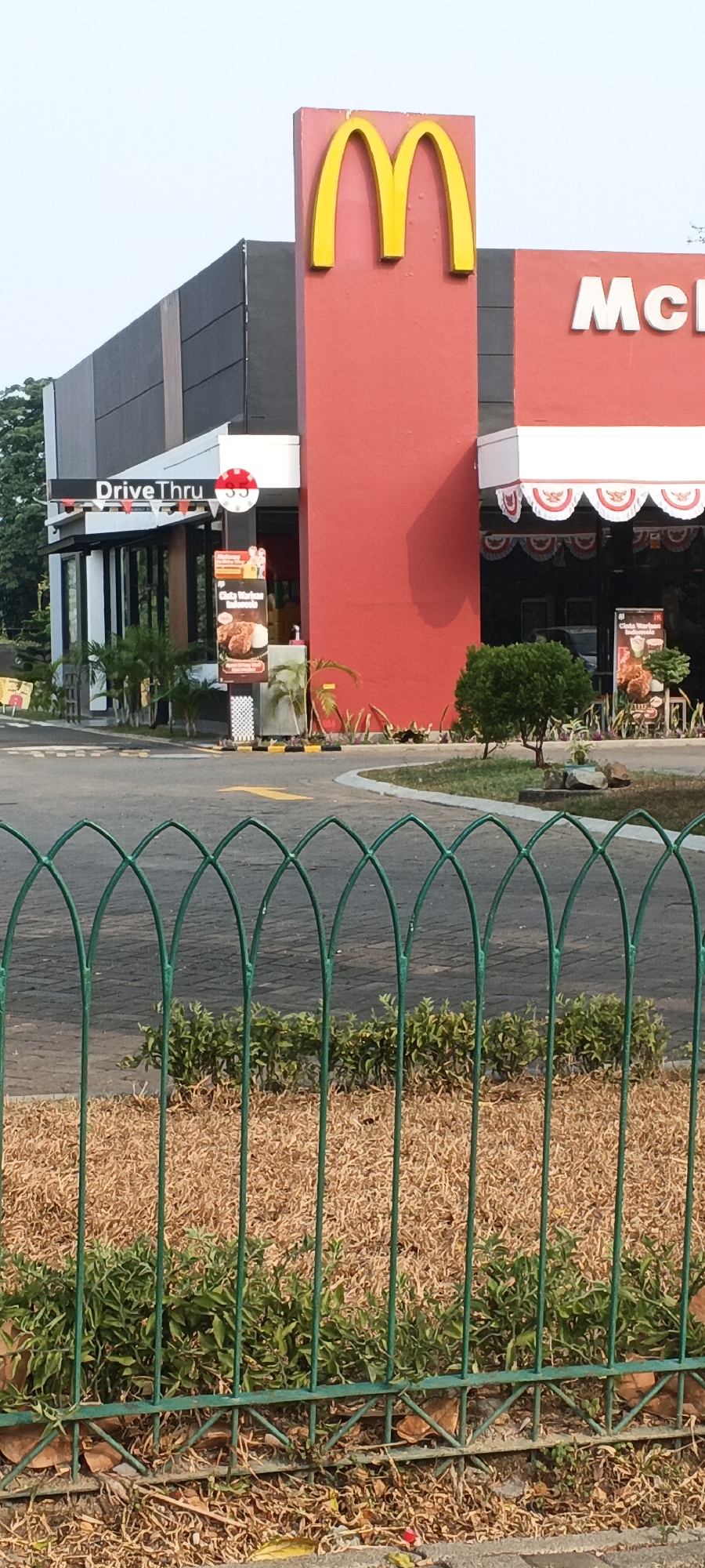 A McDonald's restaurant is visible with a prominent red facade and a large yellow 'M' logo. A drive-thru sign is also visible. The building has a modern design with dark gray and red sections. In the foreground, a green metal fence runs across the image, with dry grass and some green bushes behind it. The fence has an arched pattern. The ground is paved with asphalt, and there's a yellow arrow painted on it, indicating a lane. The sky is a pale blue.