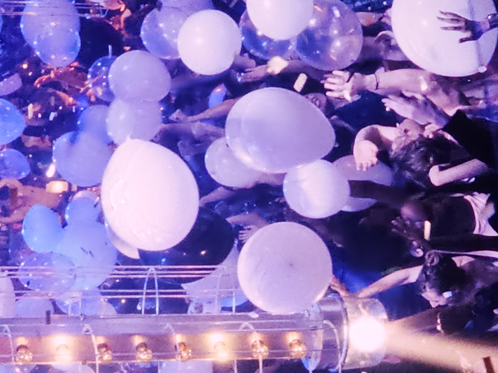 This media file captures an energetic indoor event, likely a party or club night, within a venue in Denpasar, Indonesia. The scene is dominated by a multitude of balloons and a lively crowd interacting with them.
**Subjects:** Numerous people are visible, mostly their raised arms and hands reaching upwards. Partial views of heads, shoulders, and torsos indicate a dense gathering. The people appear engaged, with various skin tones discernible. The primary objects are many balloons of varying sizes and opacities, in shades of white, translucent white, and light blue or purple. At the bottom of the frame, a metallic-looking structure, possibly part of a stage, a barrier, or equipment, is visible, equipped with several bright, warm-toned lights.
**Activity or Event:** The image depicts an active moment at a celebratory or entertainment event. People are seen reaching, touching, and interacting with the balloons, suggesting they are being released, thrown, or caught amidst the crowd. The raised hands convey excitement and participation.
**Notable Details:** The scene is illuminated by vibrant artificial lighting, creating a festive, nightclub-like atmosphere. Cool tones of blue and purple are predominant, casting an ethereal glow on the balloons and the crowd. Brighter, warm white or amber lights emanating from the structure at the bottom add dynamic highlights and lens flares to the foreground. The artificial lighting indicates an indoor setting, likely at night. The overall emotion conveyed is one of joy, excitement, and engagement. There is no visible text in the image.