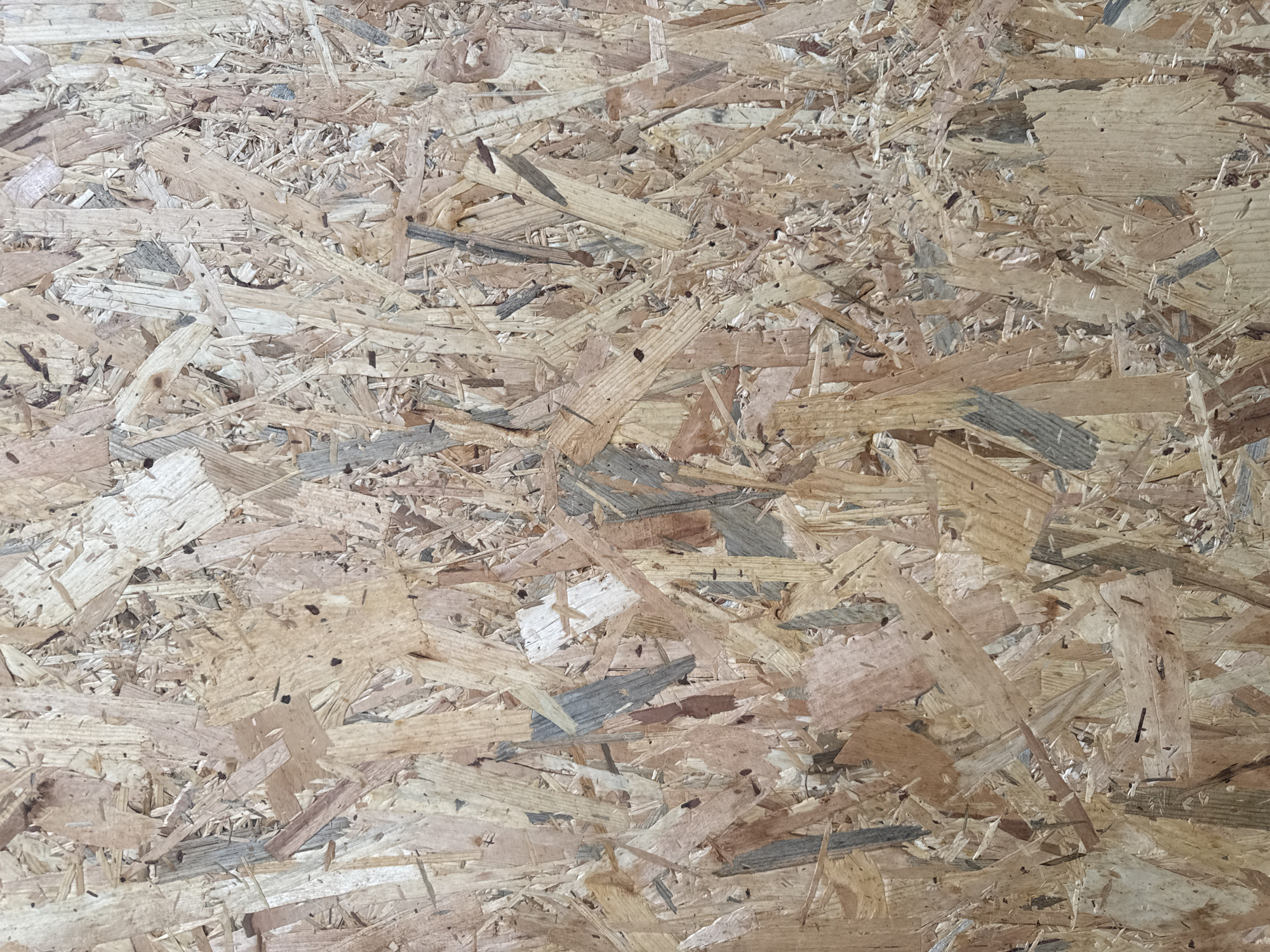 The image is a close-up, overhead shot of a particleboard surface, likely an Oriented Strand Board (OSB). The texture is rough and uneven, composed of compressed wood chips and strands of varying sizes and colors, ranging from light tan to darker brown and gray. The light casts minimal shadows, suggesting a diffuse or overhead light source. There are no discernible objects, people, or actions within the frame. The scene is devoid of any recognizable urban or natural environment, and therefore, its location in Fontevivo, Italy, cannot be determined from the image itself.