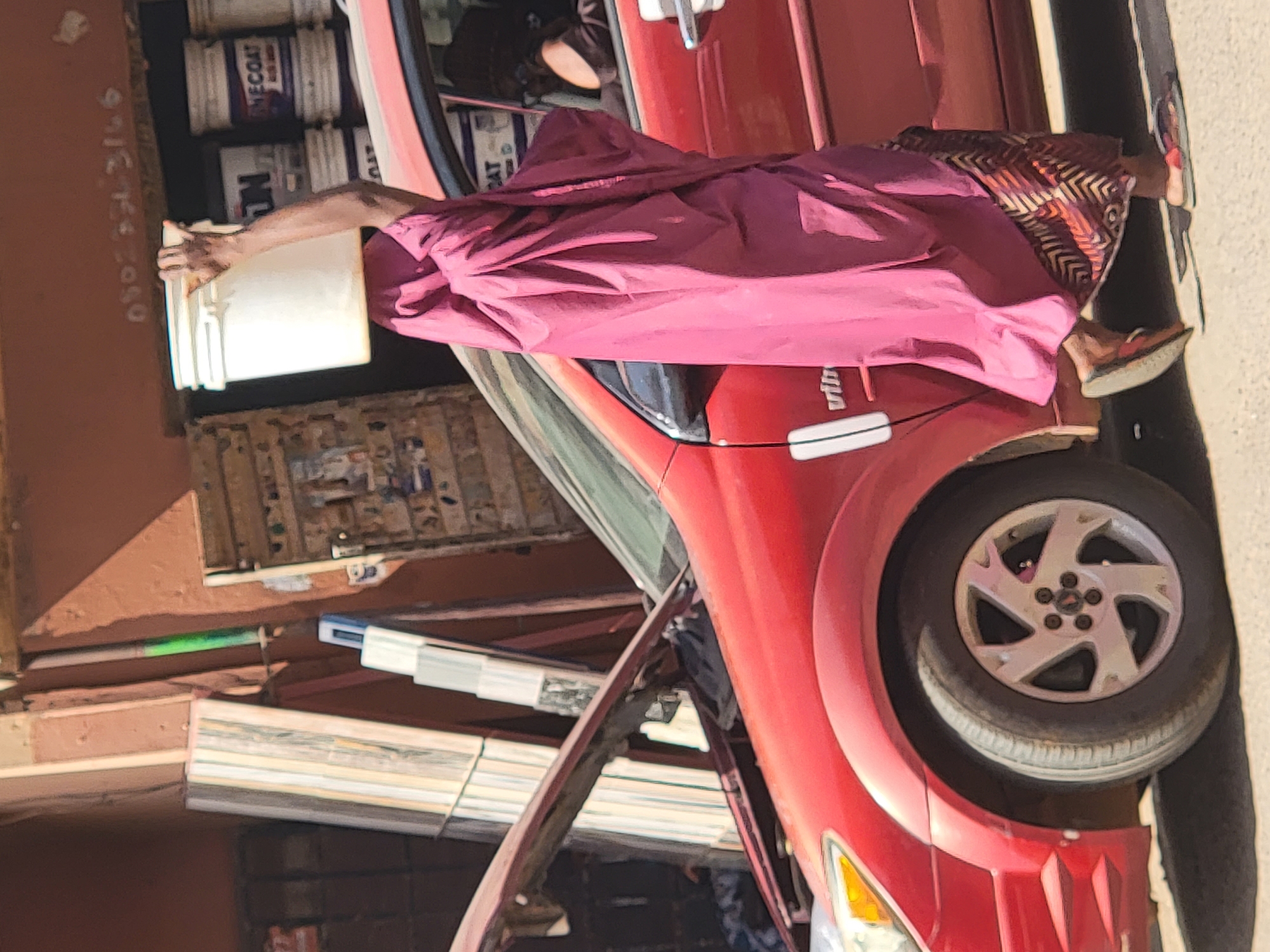 In Maiburiji, Nigeria, a person in pink clothing and patterned trousers stands next to a red car with the hood open. The individual holds a white bucket. Paint cans are visible inside a building. Bundles of materials rest on the ground near the open hood of the car.